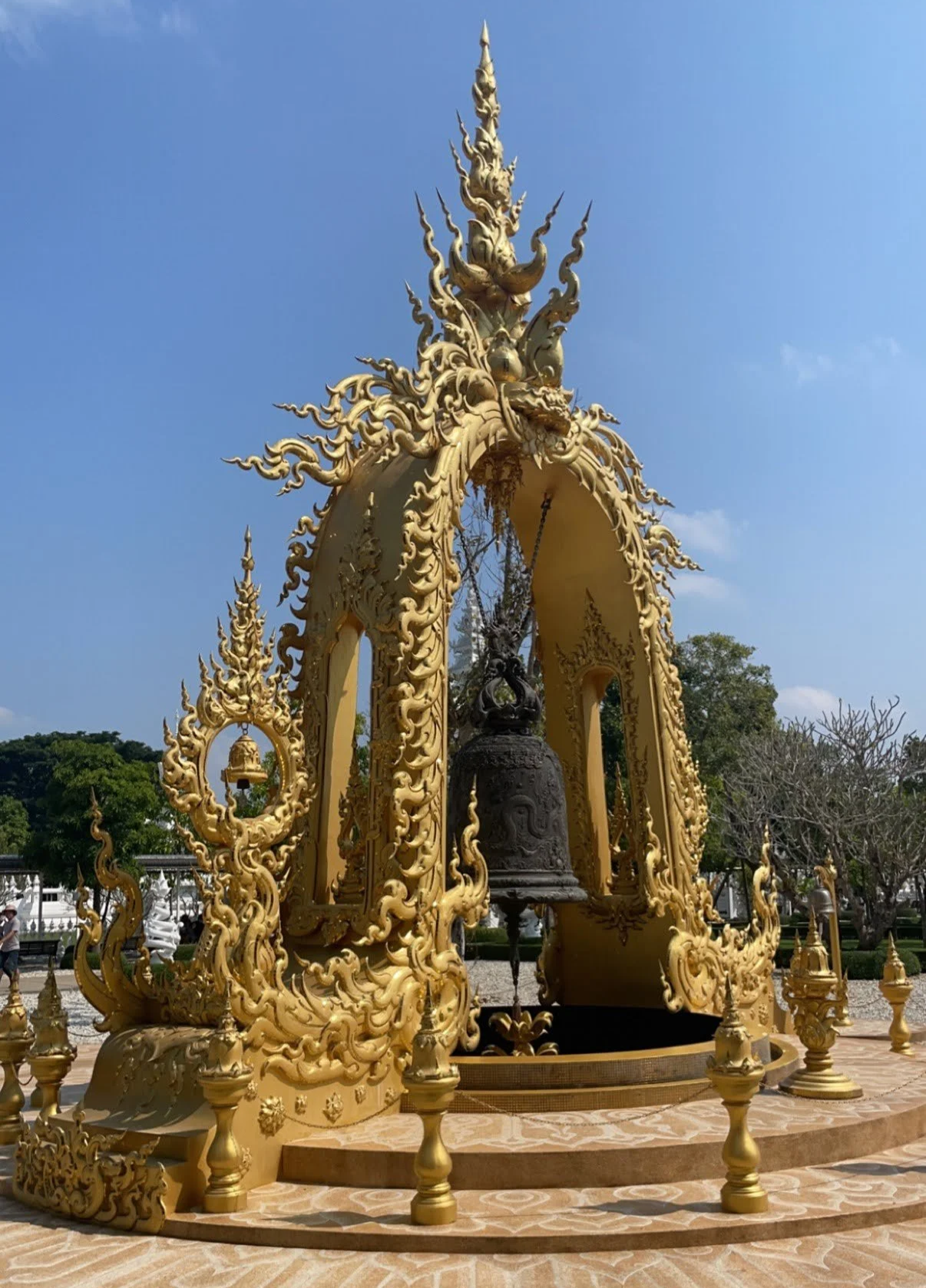 Ornate gold and black Buddhist temple bell structure with dragon carvings, set outdoors under a blue sky, surrounded by trees.