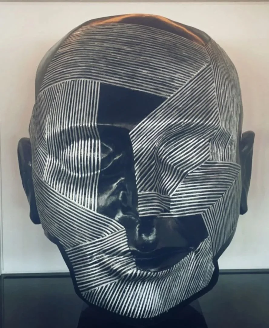 A black sculpture of a human face with white linear patterns creating fragmented segments across the face.