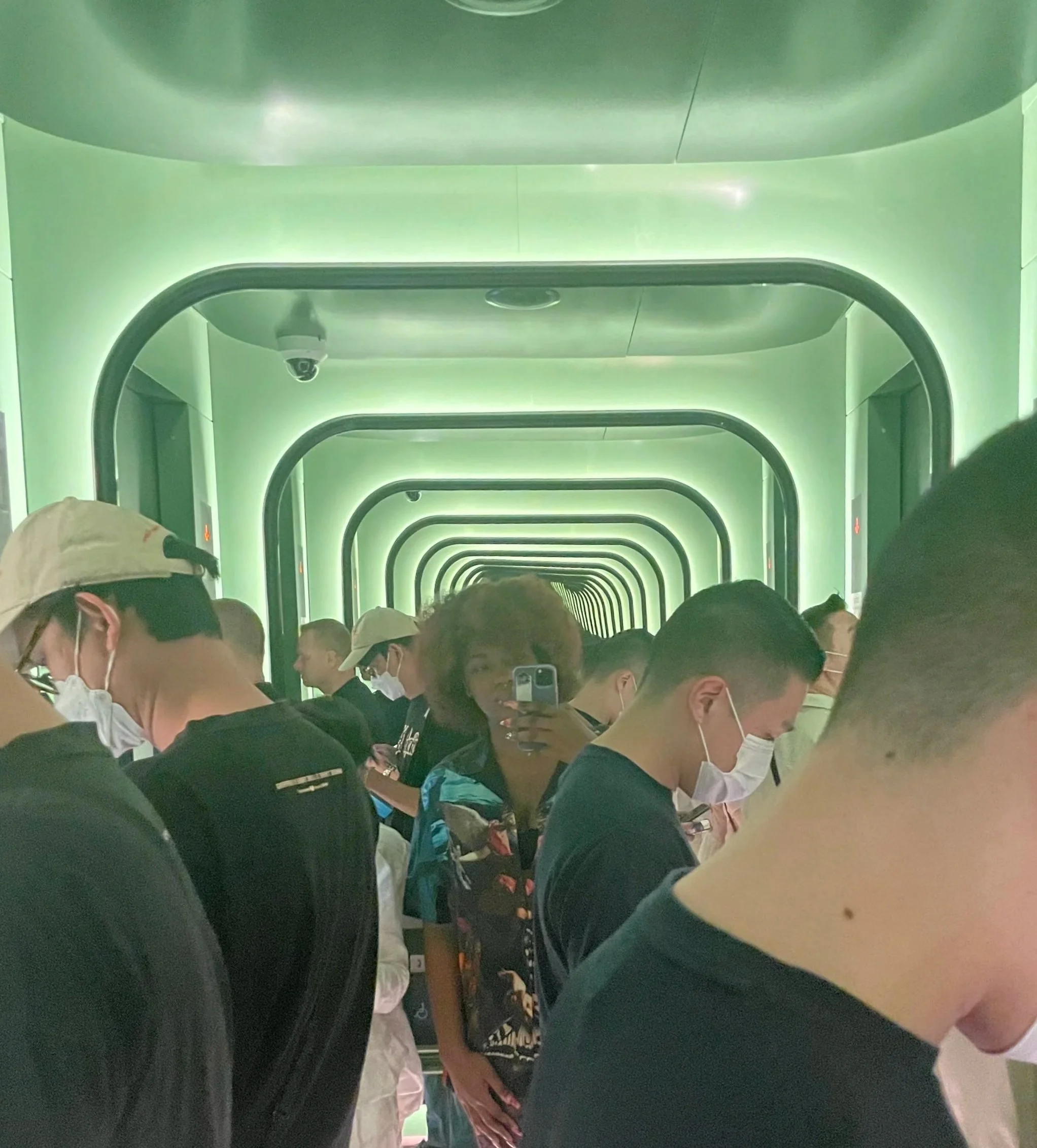 People standing in a futuristic, green-lit corridor with mirrored walls creating a tunnel effect.