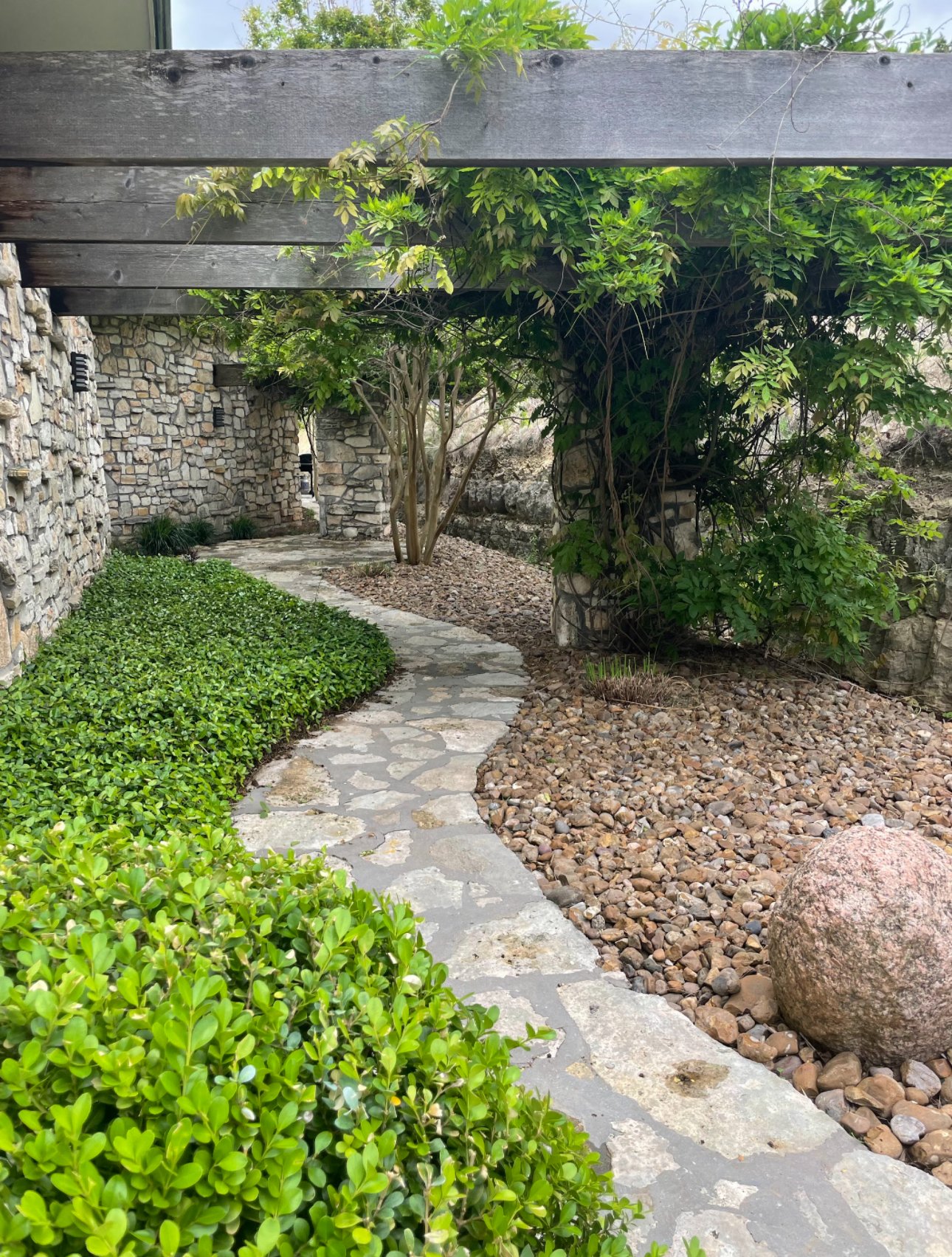 Stone pathway winding through a landscaped garden with green bushes, gravel, and climbing vines under a wooden pergola.