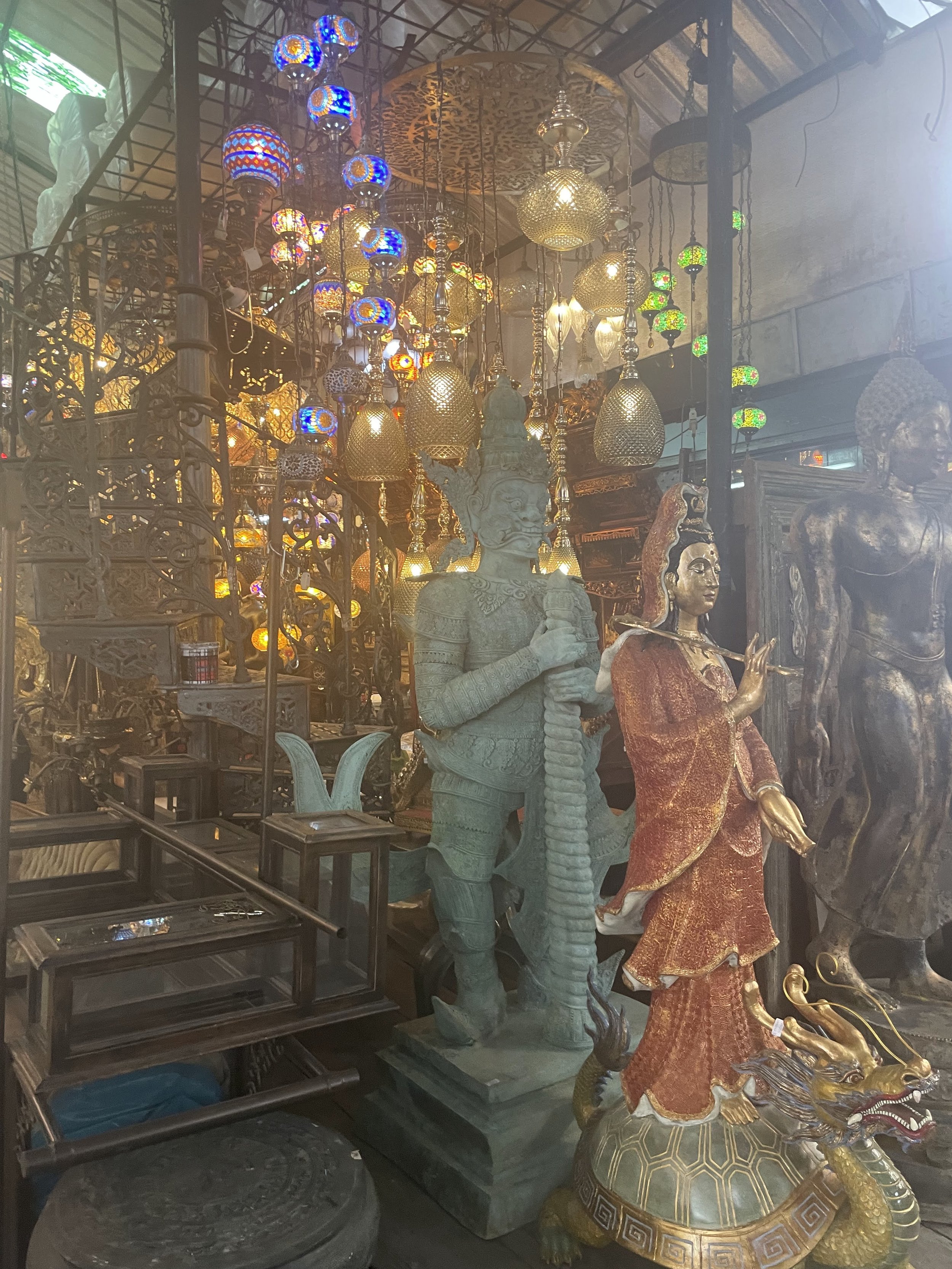 Various intricately designed statues of mythological figures and deities, with colorful hanging lanterns and lamps in the background, in an antique shop or display setting.