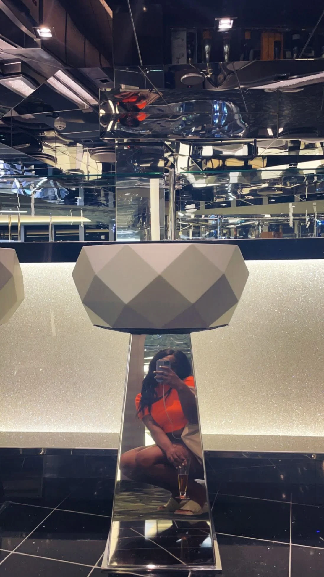 Mirror with a woman taking a photo, squatting with a drink, in a modern bar or restaurant with shiny, reflective decor and geometric lighting fixtures.
