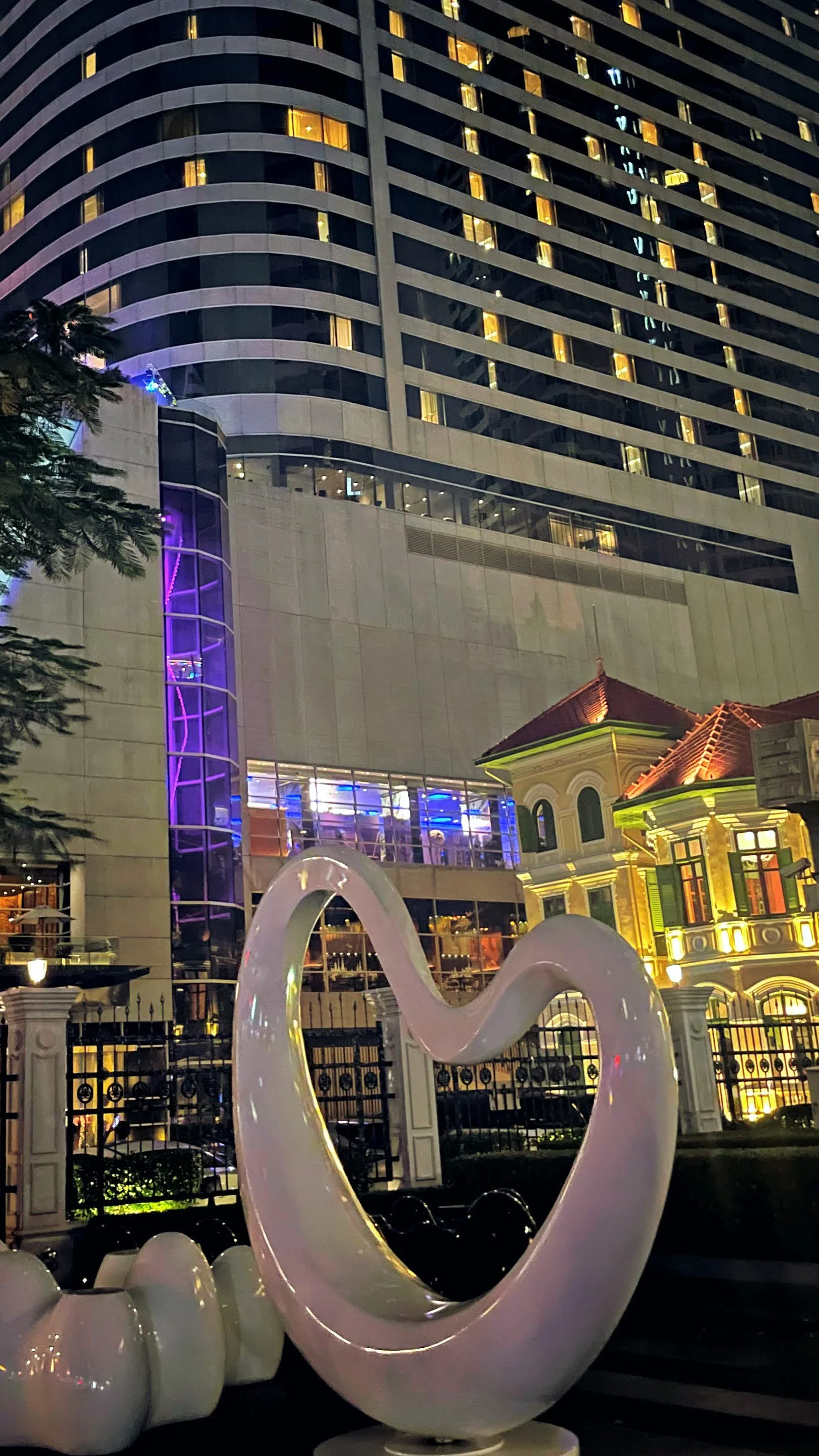 A modern high-rise building decorated with colorful lights, with a small ornate house in front and a large white sculpture of a heart shape in the foreground.