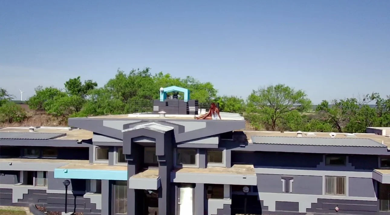 A modern multi-story apartment building with a flat roof, balconies, and large windows, surrounded by green trees and open sky.