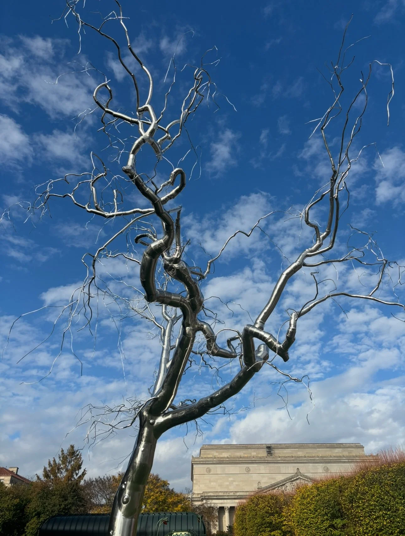 A metallic, abstract, twisted tree sculpture with numerous curving branches set against a partly cloudy blue sky, with a large historic building and some greenery in the background.