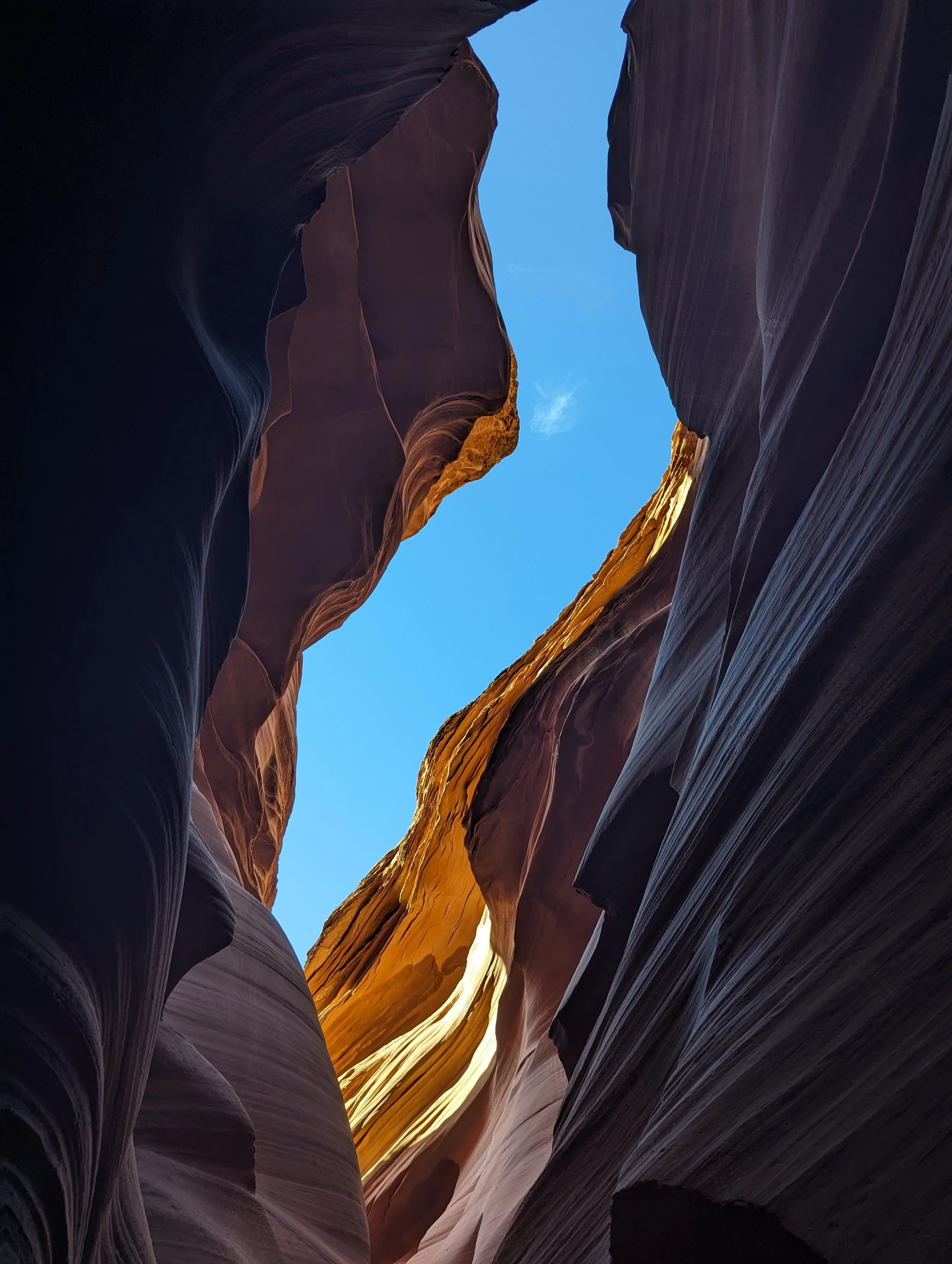 View of a narrow slot canyon with smooth, layered rock walls in shades of dark purple, brown, and orange, and a bright blue sky visible at the top.