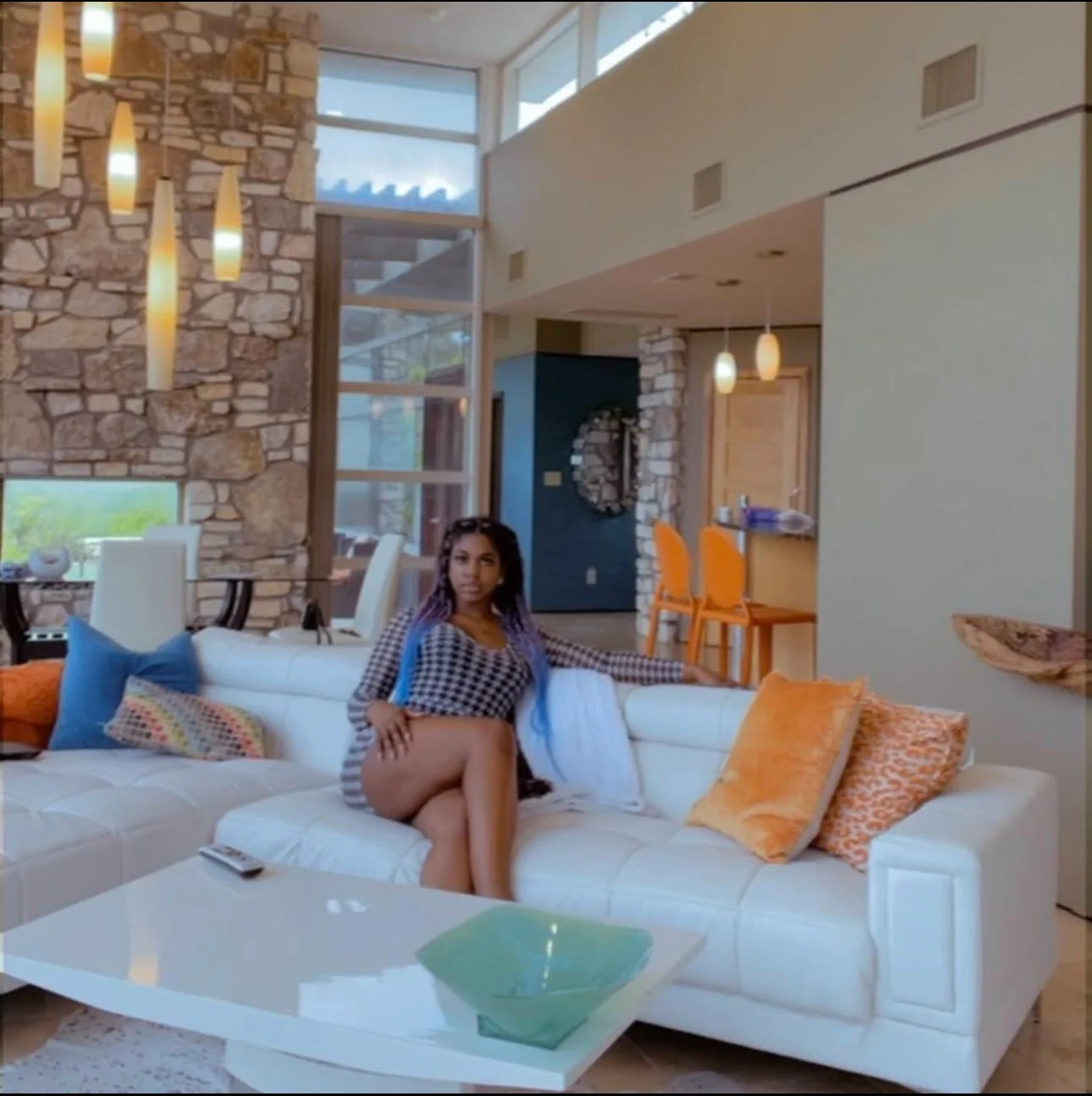 A woman with purple braided hair sitting on a white sofa in a modern, well-lit living room with stone accents, colorful pillows, and large windows.
