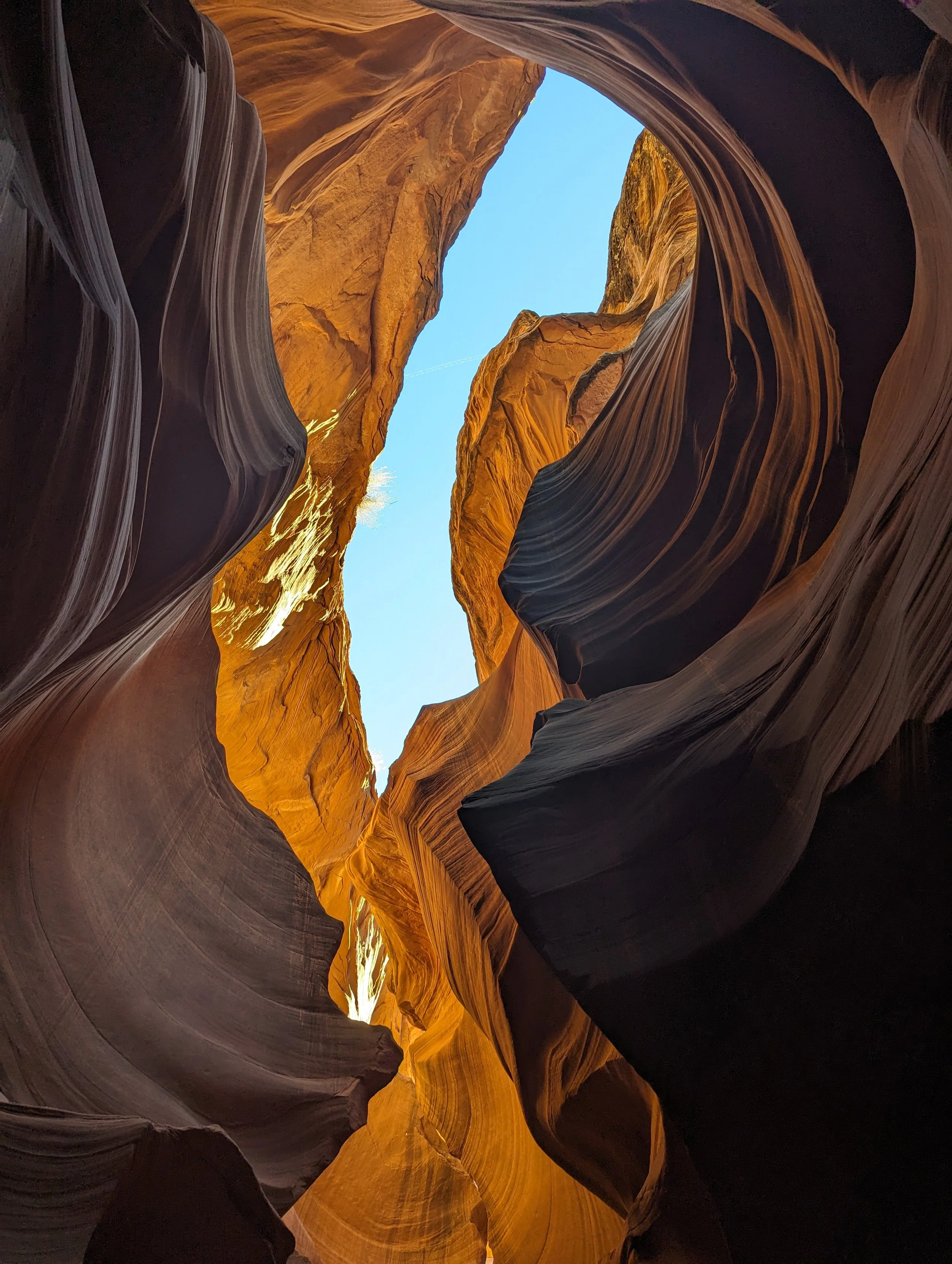 View of a narrow slot canyon with smooth, layered rock walls and a bright blue sky overhead.
