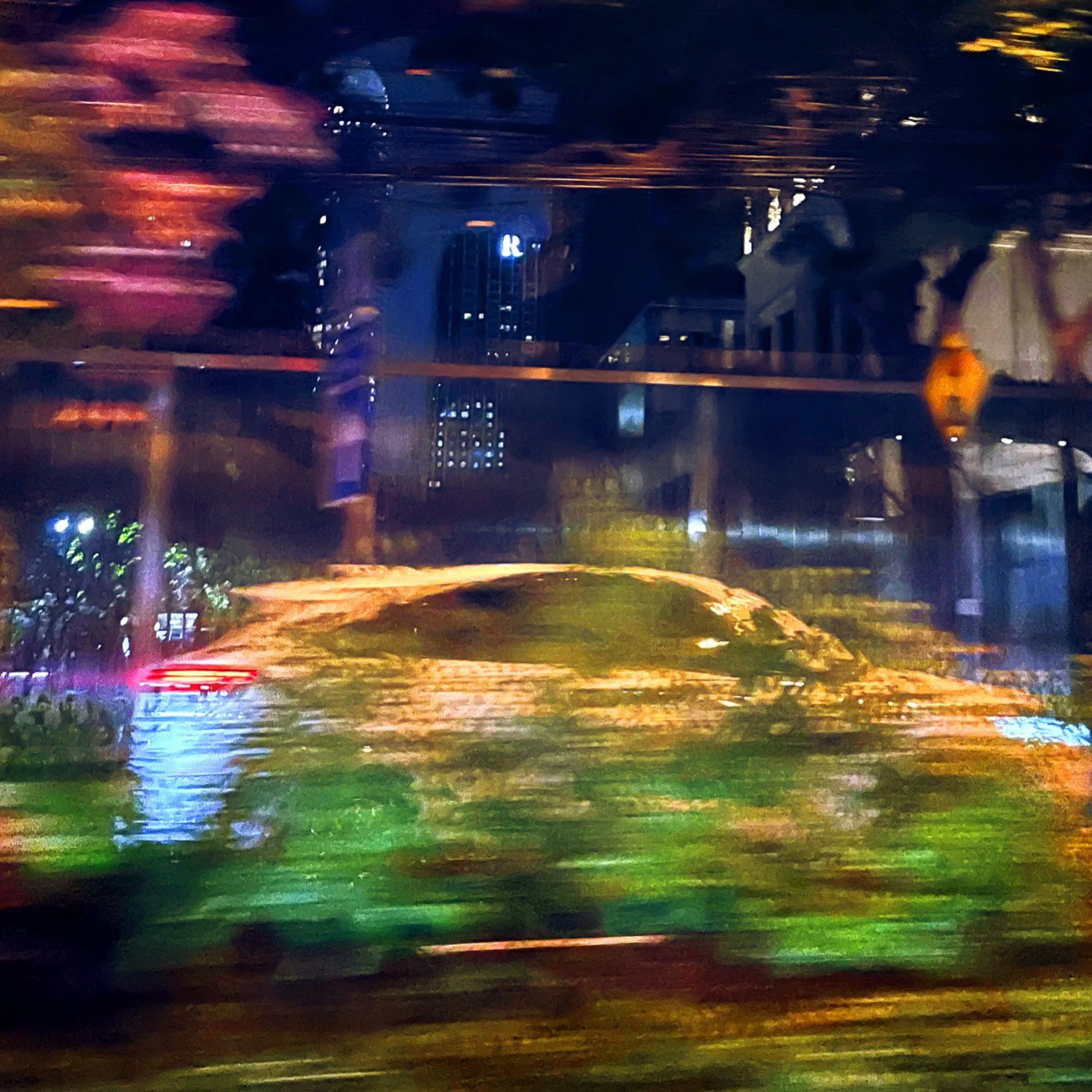 Blurred city scene at night seen through a moving vehicle window with streaks of light and indistinct buildings and trees.