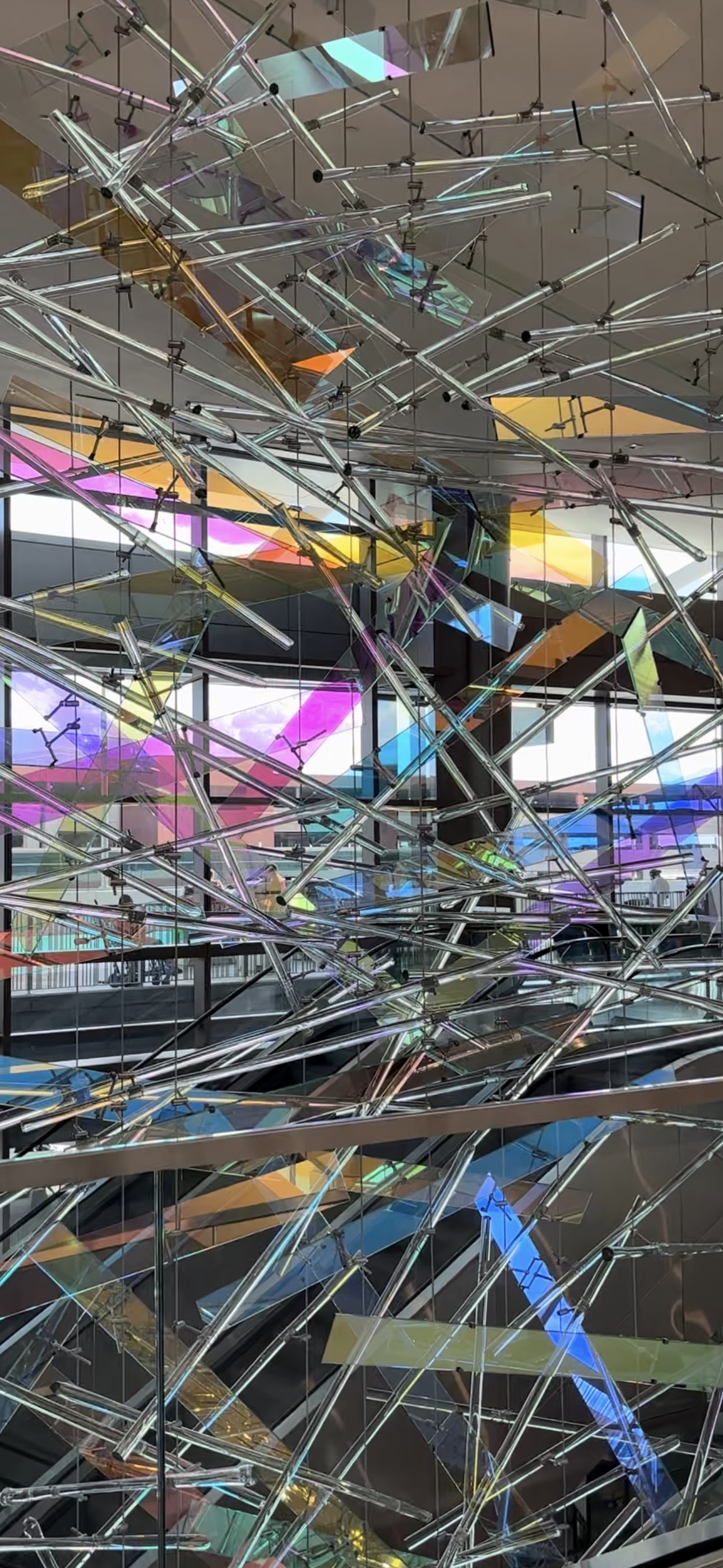 A colorful, abstract sculpture made of irregular, transparent, and reflective acrylic or glass pieces, suspended from the ceiling with black wires in a modern indoor space with large windows and multicolored light reflections.