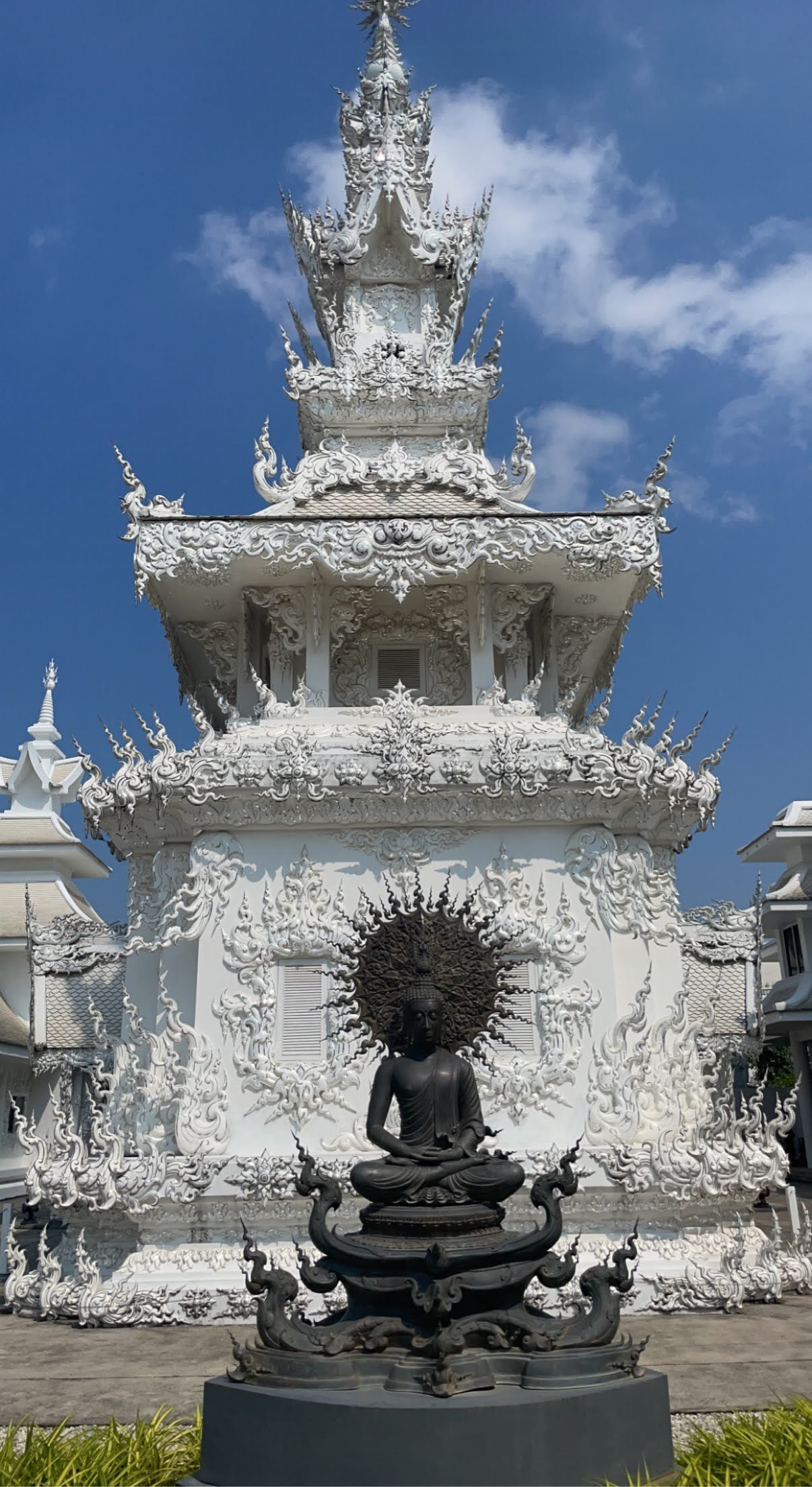 A white, ornate, multi-tiered temple structure with intricate carvings, featuring a Buddha statue seated in meditation in front, against a bright blue sky.