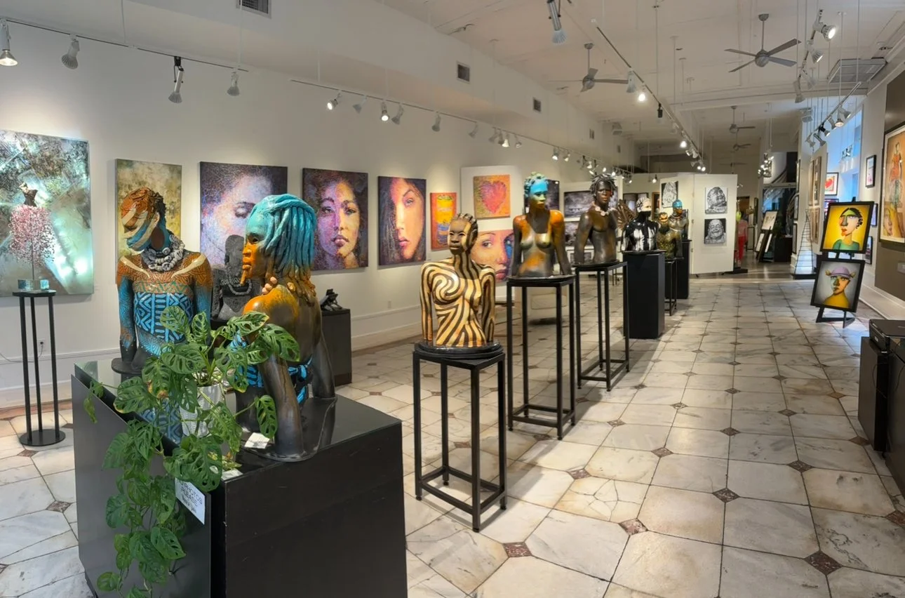 Art gallery with colorful paintings on the walls and several unique, artistic bust sculptures on black pedestals, including one with blue dreadlocks, in a well-lit space with tiled floor.