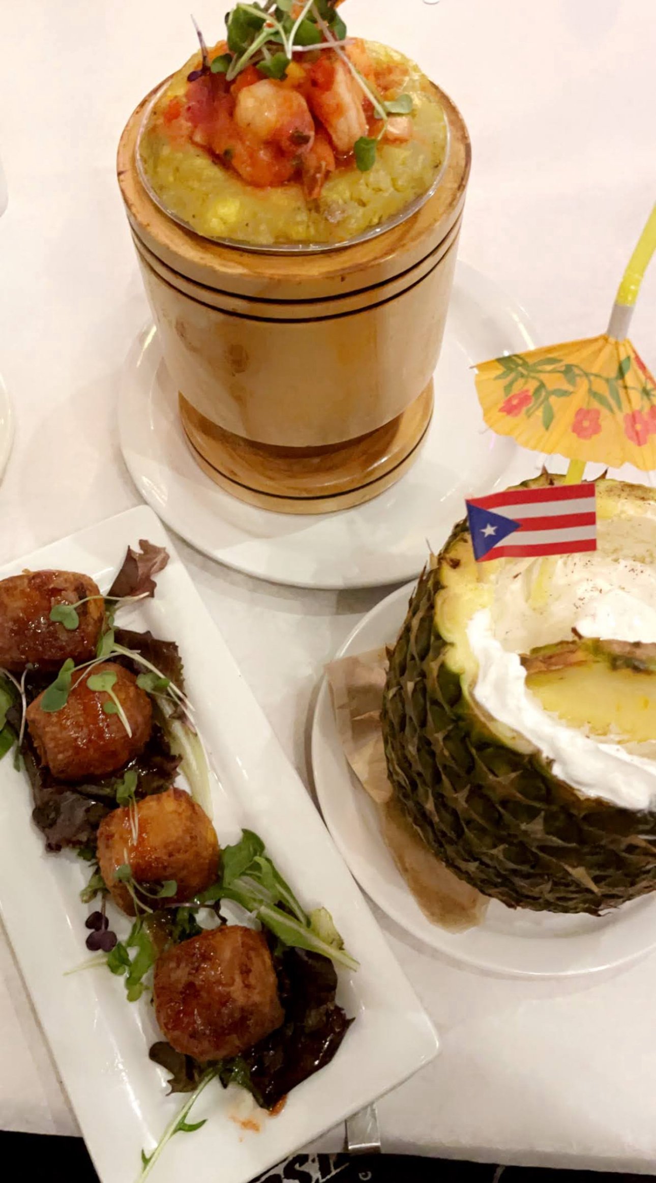 An assortment of Puerto Rican dishes includes a wooden bowl of yellow rice topped with stewed chicken, a pineapple half with whipped cream and a Puerto Rican flag toothpick, and a rectangular plate of croquettes garnished with microgreens.
