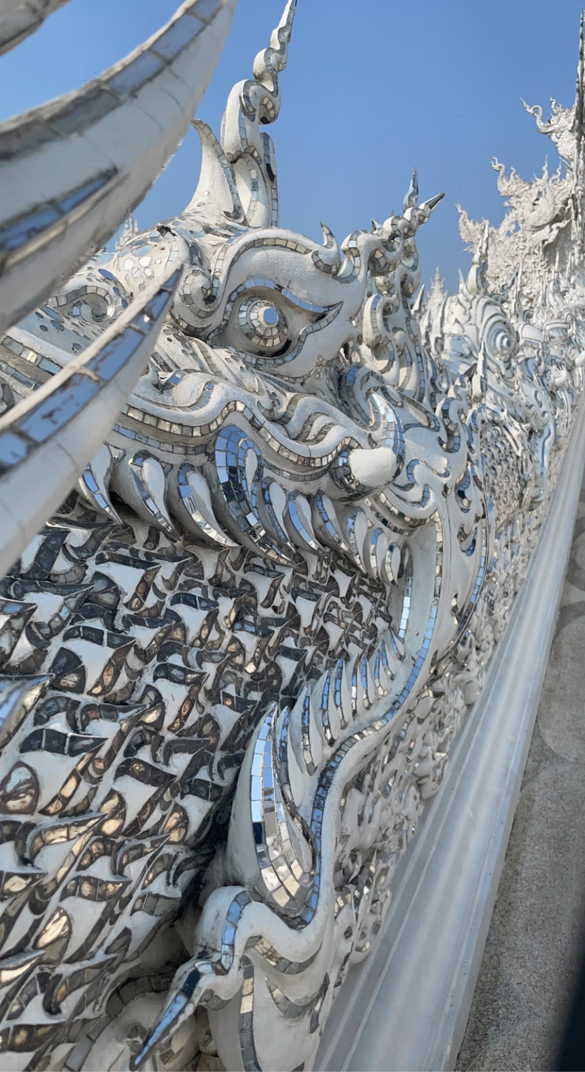  Ornate white sculpture of a mythical creature with intricate details and mirror-like reflective surfaces, set against a blue sky.