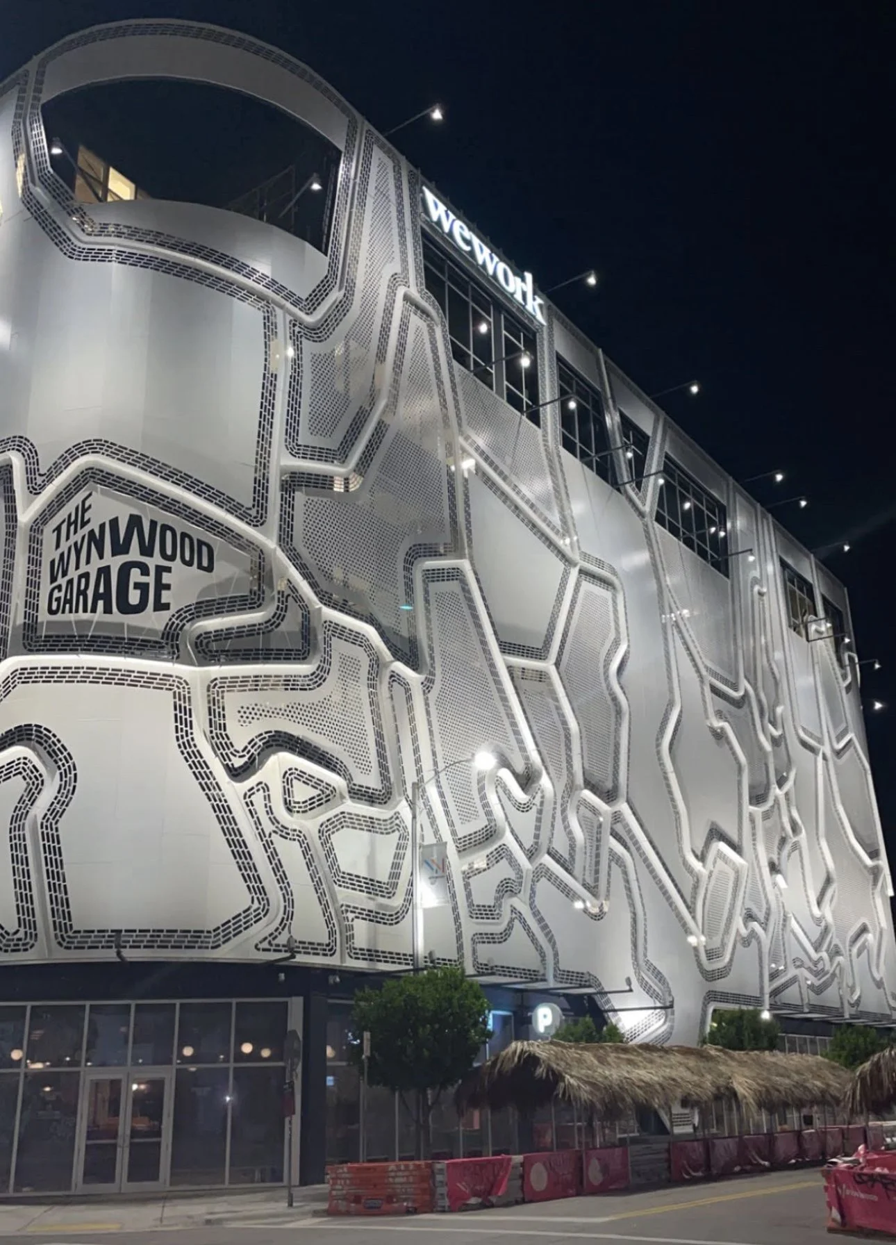 Night view of a modern multi-story building with artistic metal facade, illuminated signs for 'WeWork' and 'The Wynwood Garage', and street-level storefronts with small trees and thatched roof awnings.