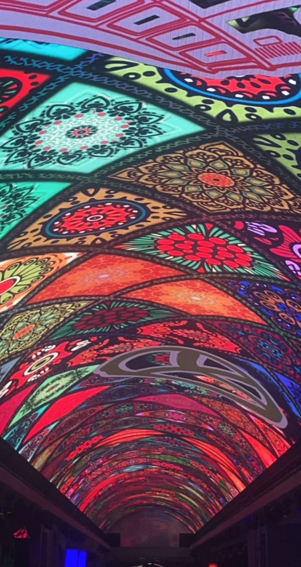 Colorful, illuminated architectural ceiling with intricate, patterned designs in various bright colors.