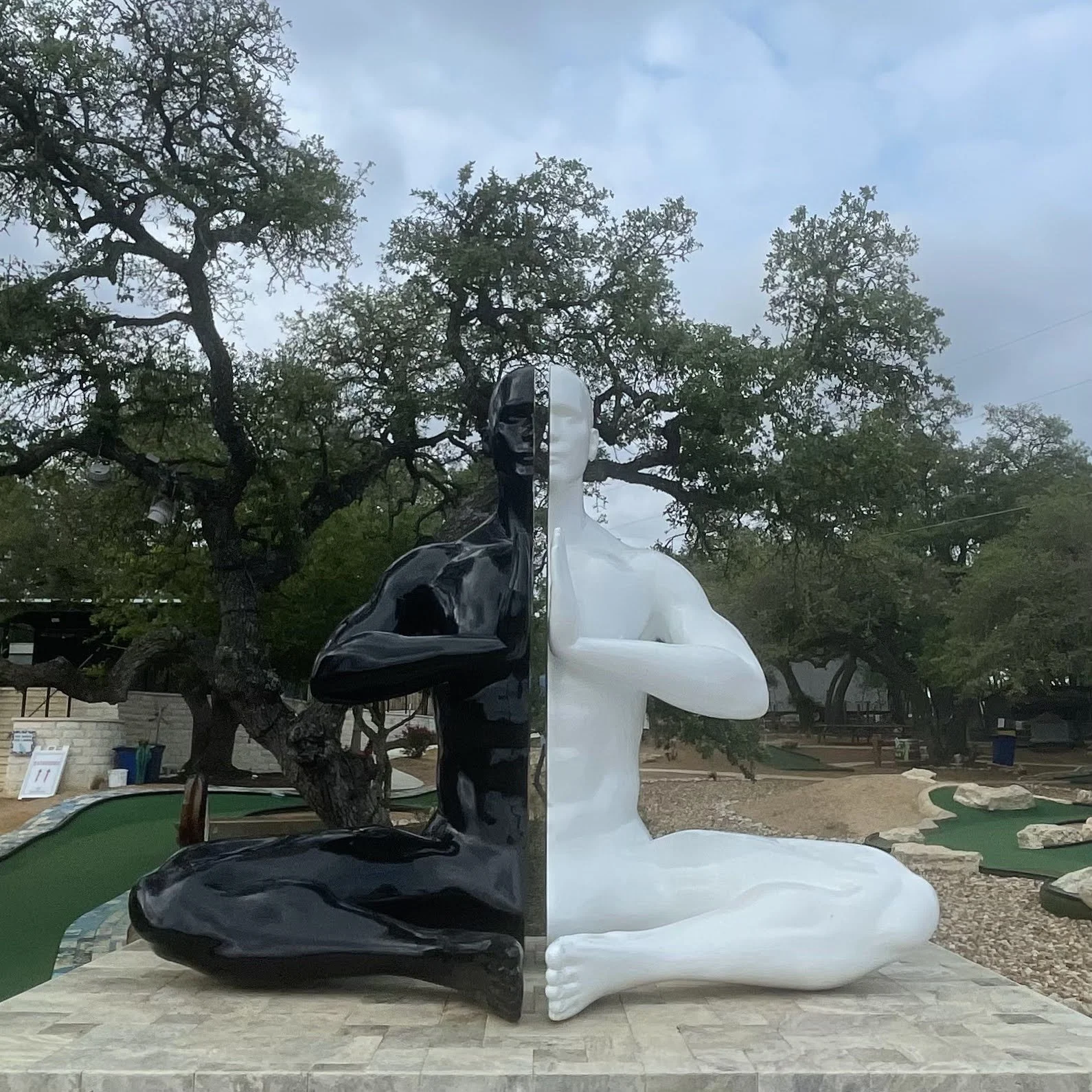 A sculpture of a human figure divided into two halves, one black and one white, sitting with crossed legs and arms crossed in front of the chest, outdoors with trees and a mini-golf course in the background.