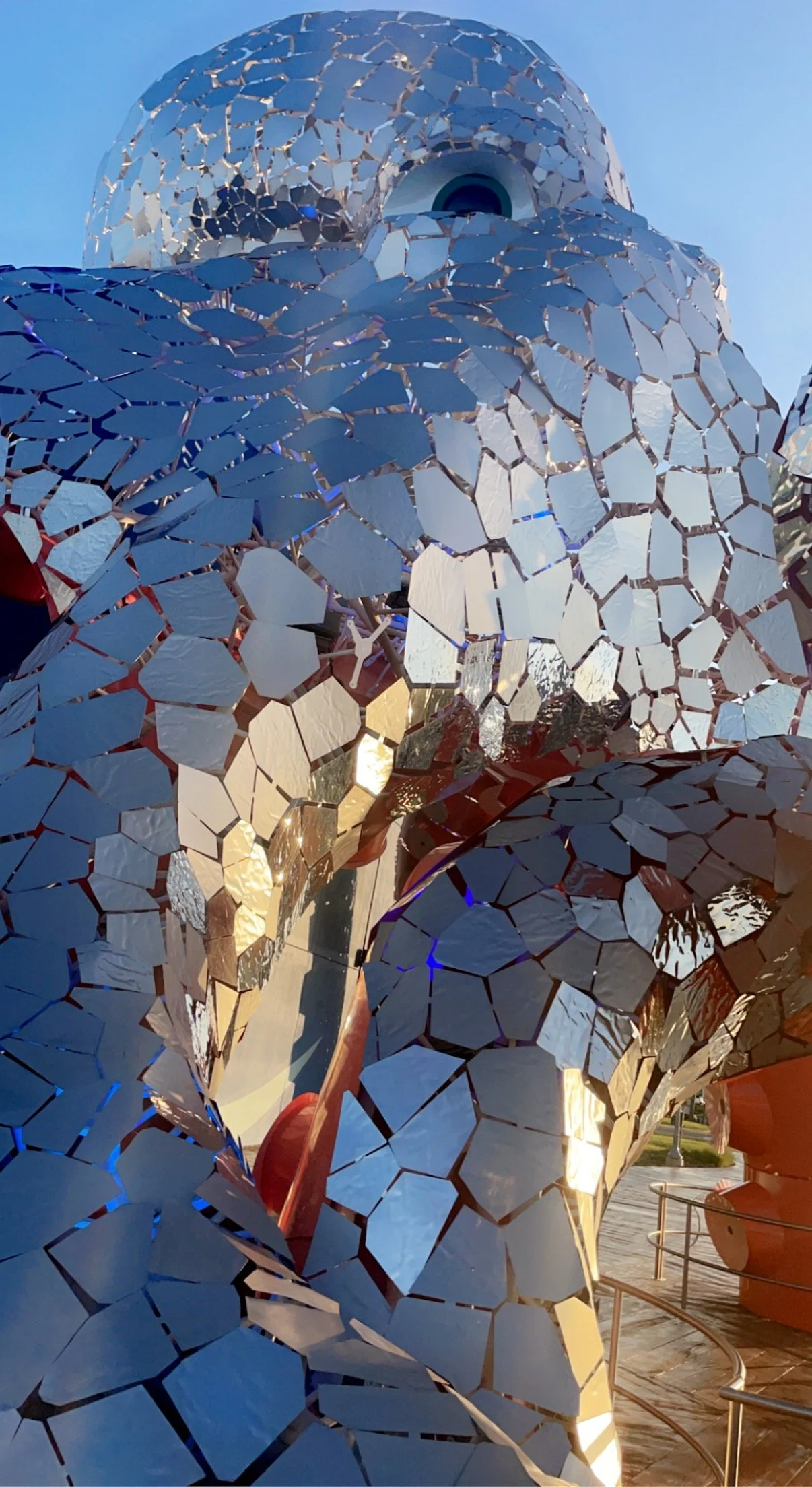 Close-up of a modern sculpture made of mirrored, polygon-shaped tiles reflecting sunlight and surrounding scenery, part of an outdoor installation.
