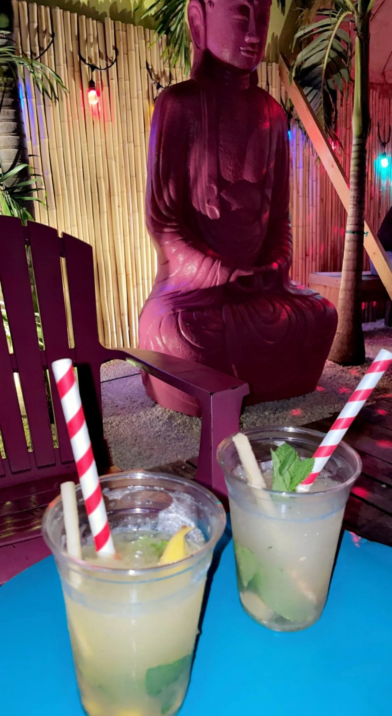 Two glasses of lemonade with mint and lemon slices, set on a blue table in front of a wooden bench and seating area with a Buddha statue and tropical plants, decorated with colorful string lights.