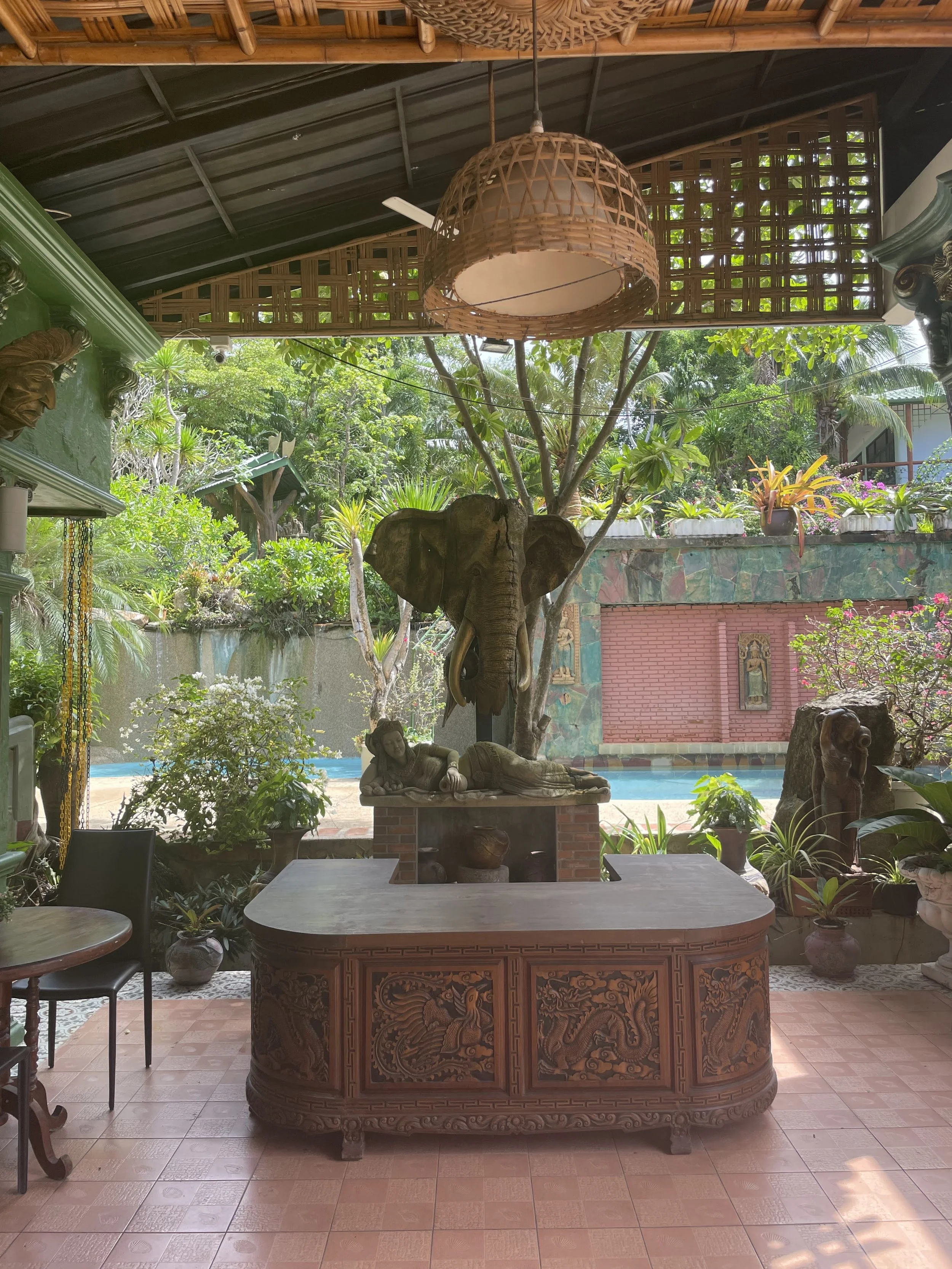 Interior view of a room with wooden furniture, decorative sculptures, and potted plants, featuring an outdoor garden with trees and plants visible through open windows.