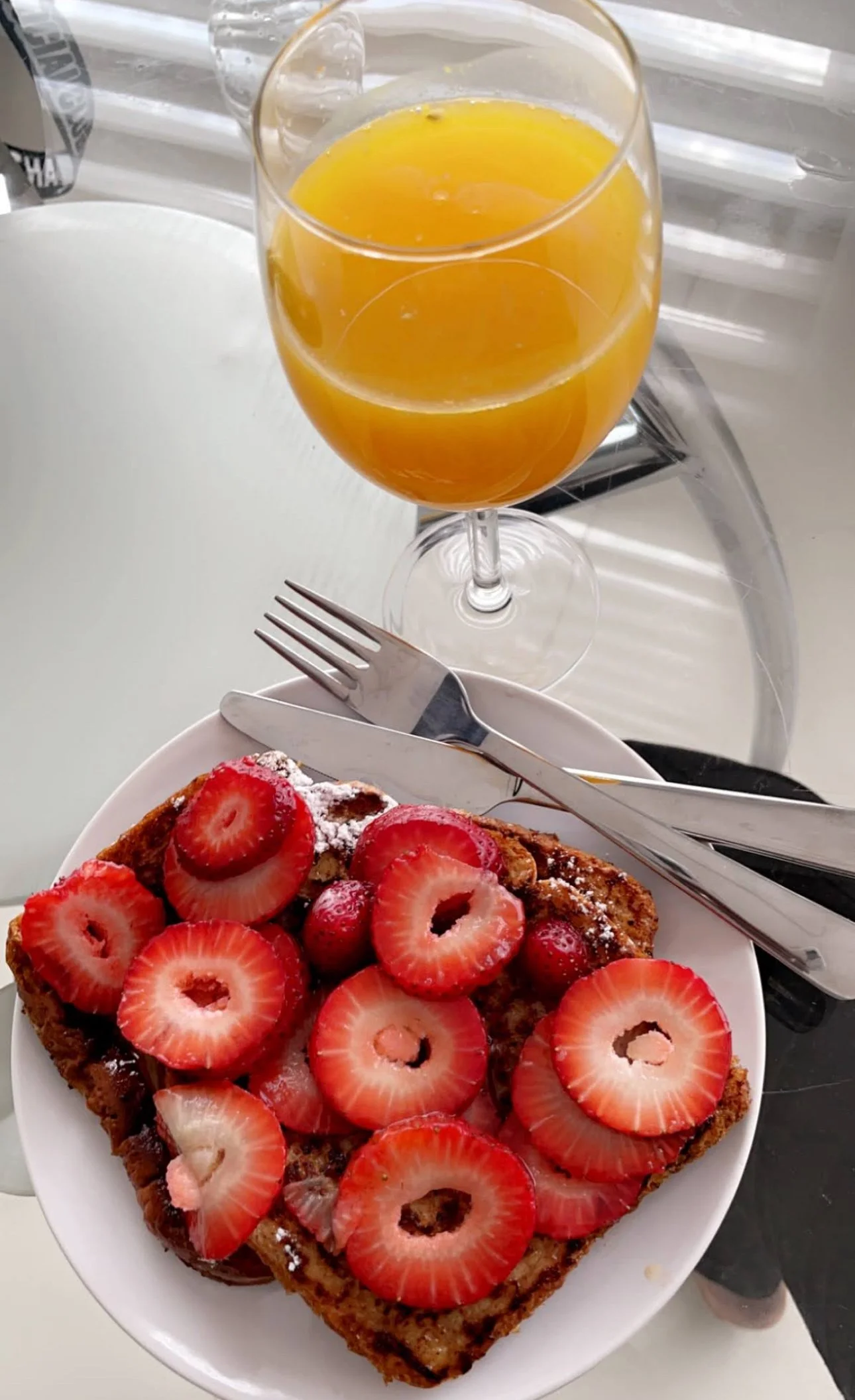 A breakfast plate with strawberry-topped French toast, a fork and knife, a glass of orange juice, and a white plate in the background.