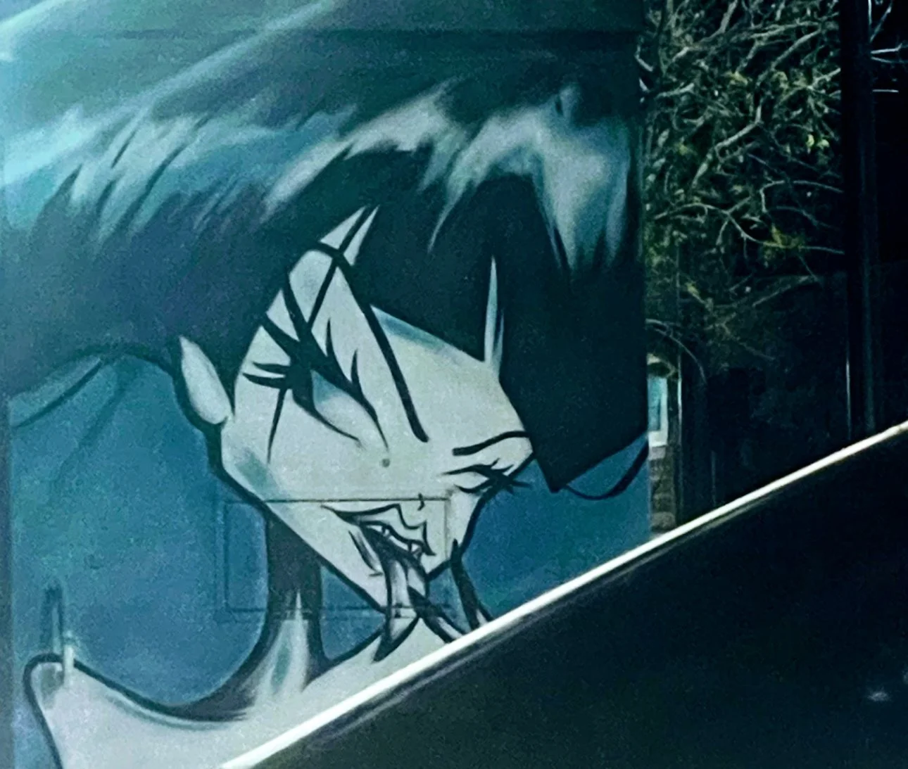 Street art mural of a character with spiky hair, angry expression, sharp teeth, and shadows, painted on a blue wall.