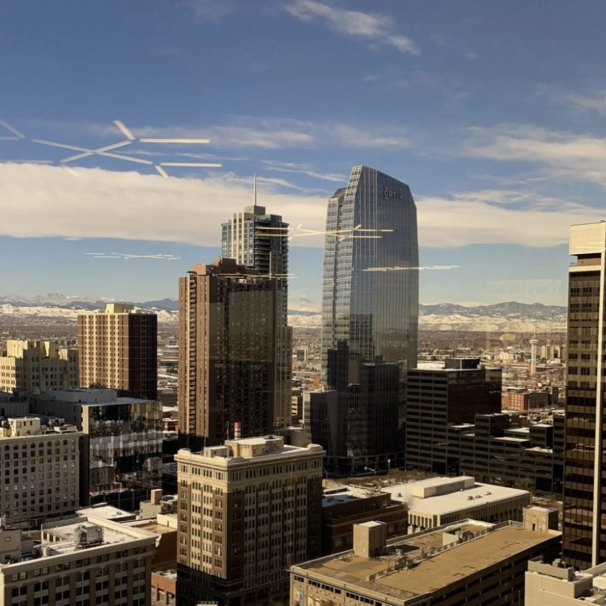 City skyline with tall buildings, snow-capped mountains in the background, and aircraft contrails in the sky.