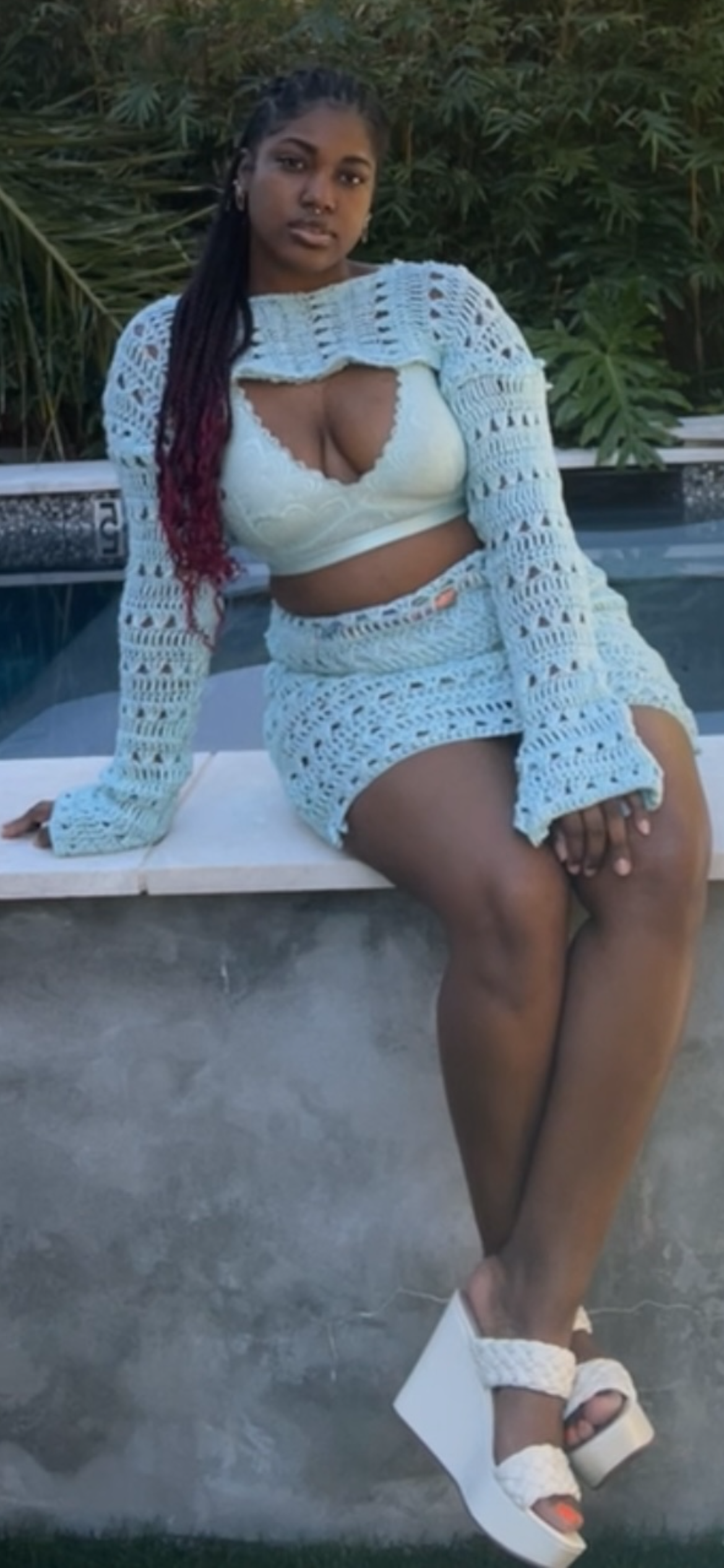 A woman sitting by a pool, wearing a white lace top and skirt with a crochet jacket, white high-heeled sandals, and sporting long braids with red highlights.