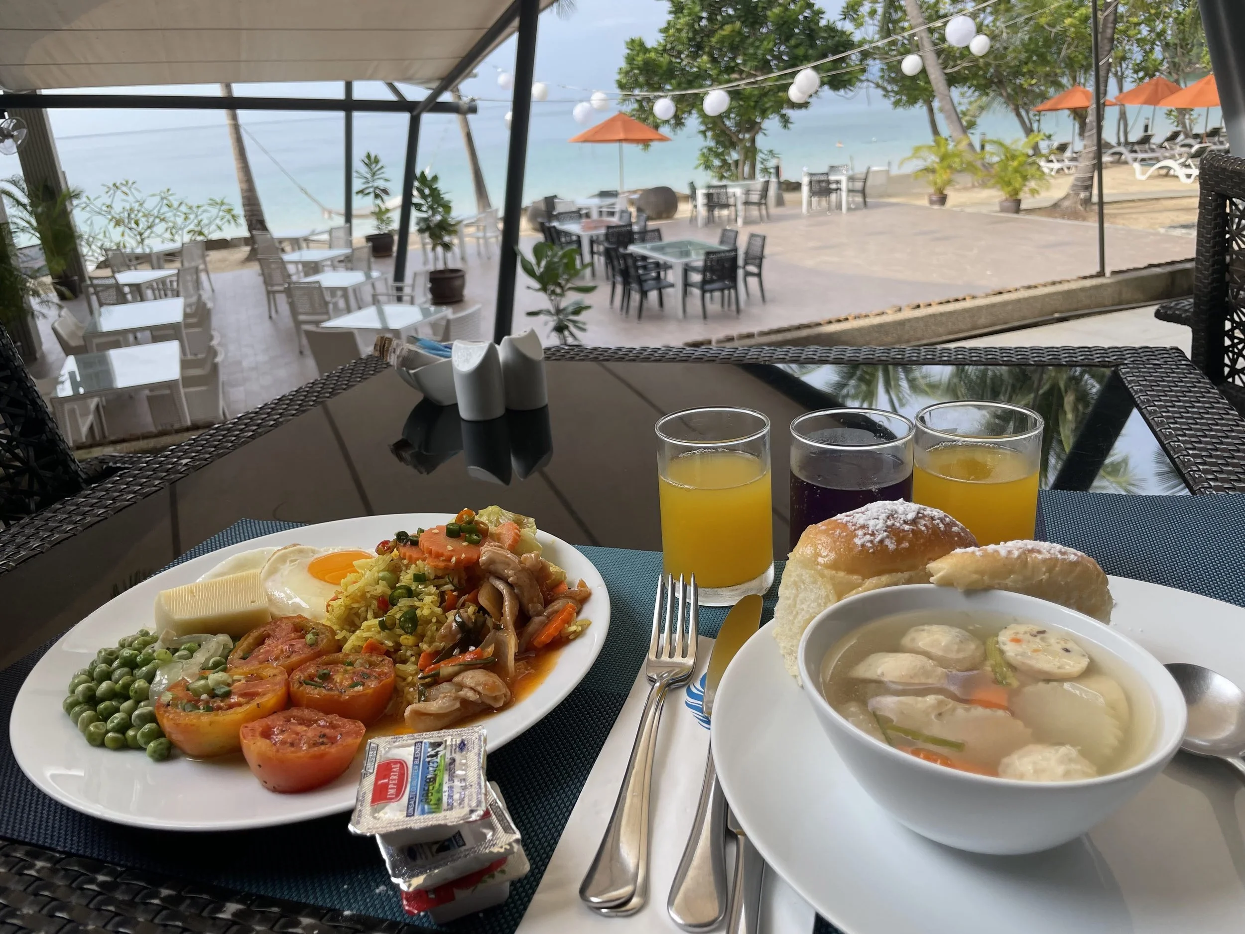 A table with breakfast food and drinks overlooking a beachside patio with umbrellas and chairs, sea view, and string lights.