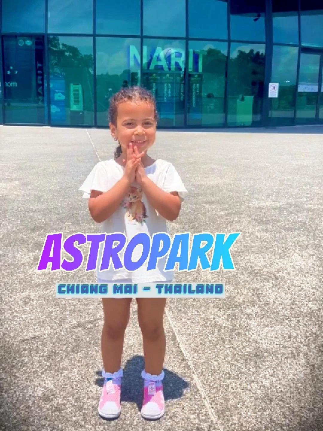 Una bambina sorridente con abbigliamento casual in piedi all'aperto davanti a un edificio con vetri, con un cartello che dice "ASTROPARK" e "Chiang Mai - Thailand".