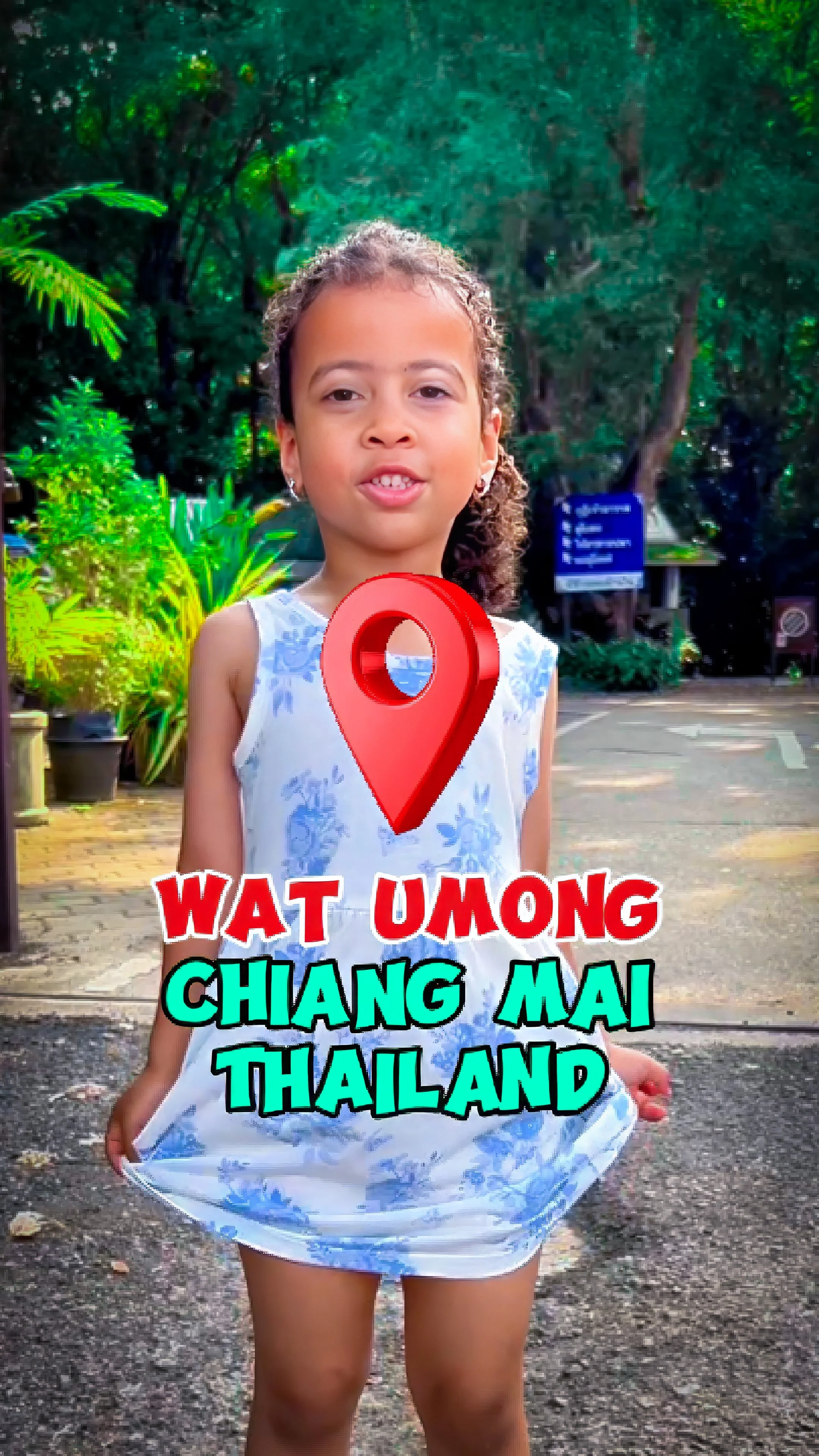 Una giovane ragazza davanti a un ambiente naturale con vegetazione verde, indossa un vestito bianco e soffia un bacio. Sulla foto c'è un segnaposto rosso con un'icona di posizione e il testo in inglese e italiano 'What among Chiang Mai Thailand'.