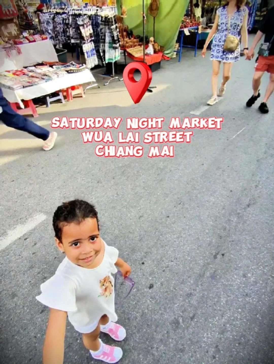 Bambina sorridente con capelli riccioli che si guarda e si prende una selfie in una strada del mercato notturno di Wua Lai Street a Chiang Mai, con bancarelle che vendono vari prodotti e alcune persone che passeggiano.