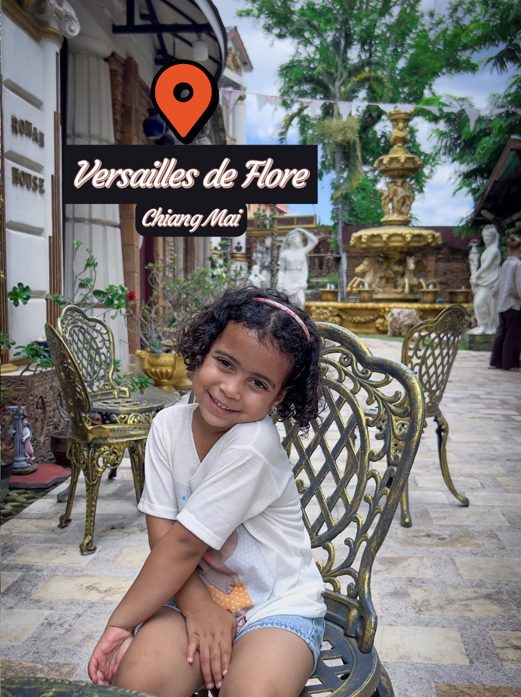 Una bambina sorridente seduta su una sedia di metallo in un cortile decorato con statue e fontane, con un edificio antico sullo sfondo, in Chiang Mai, Thailandia.