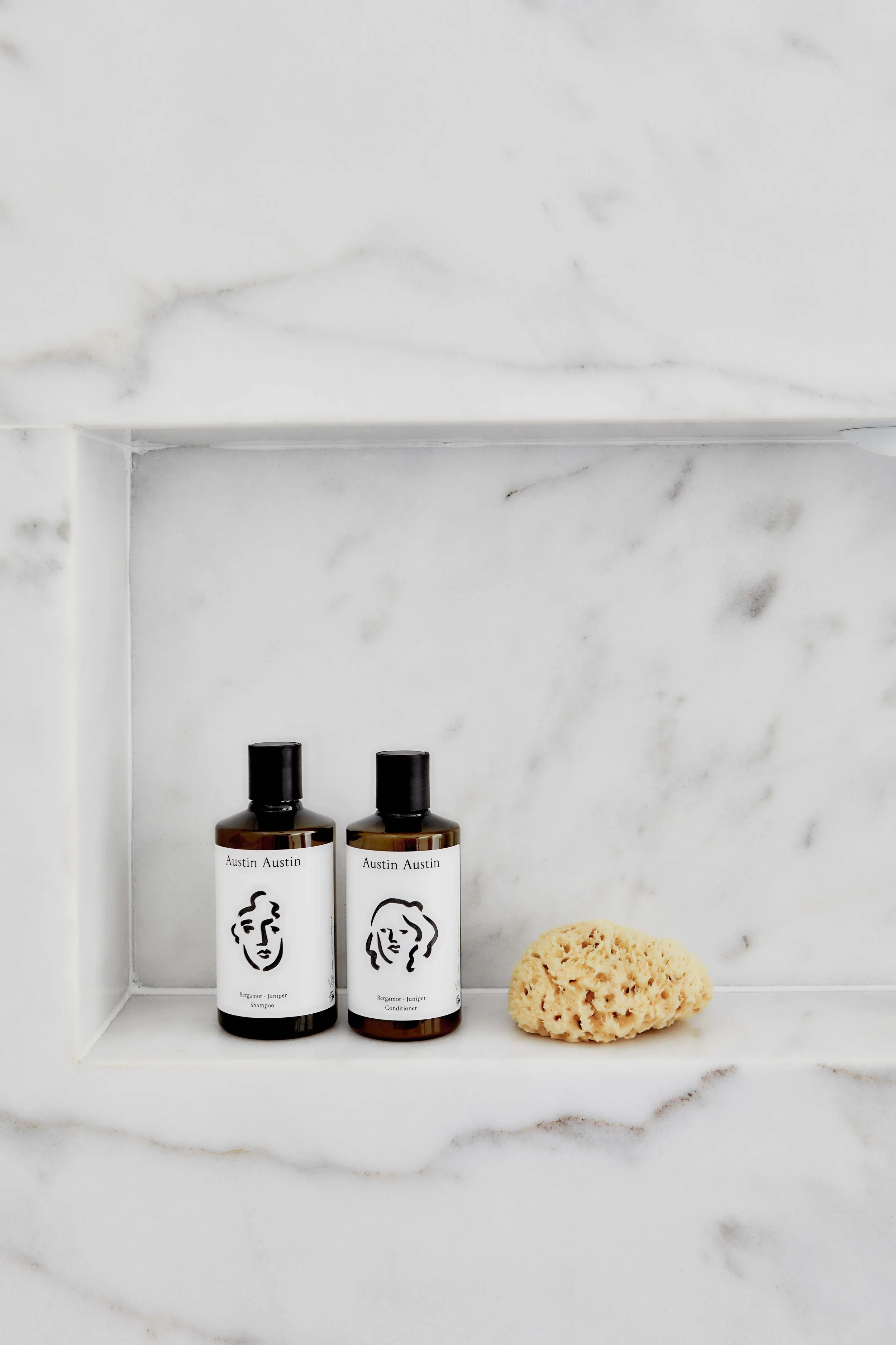 Two brown bottles labeled Austin Austin, one shampoo and one conditioner, a natural sponge on a marble shelf.
