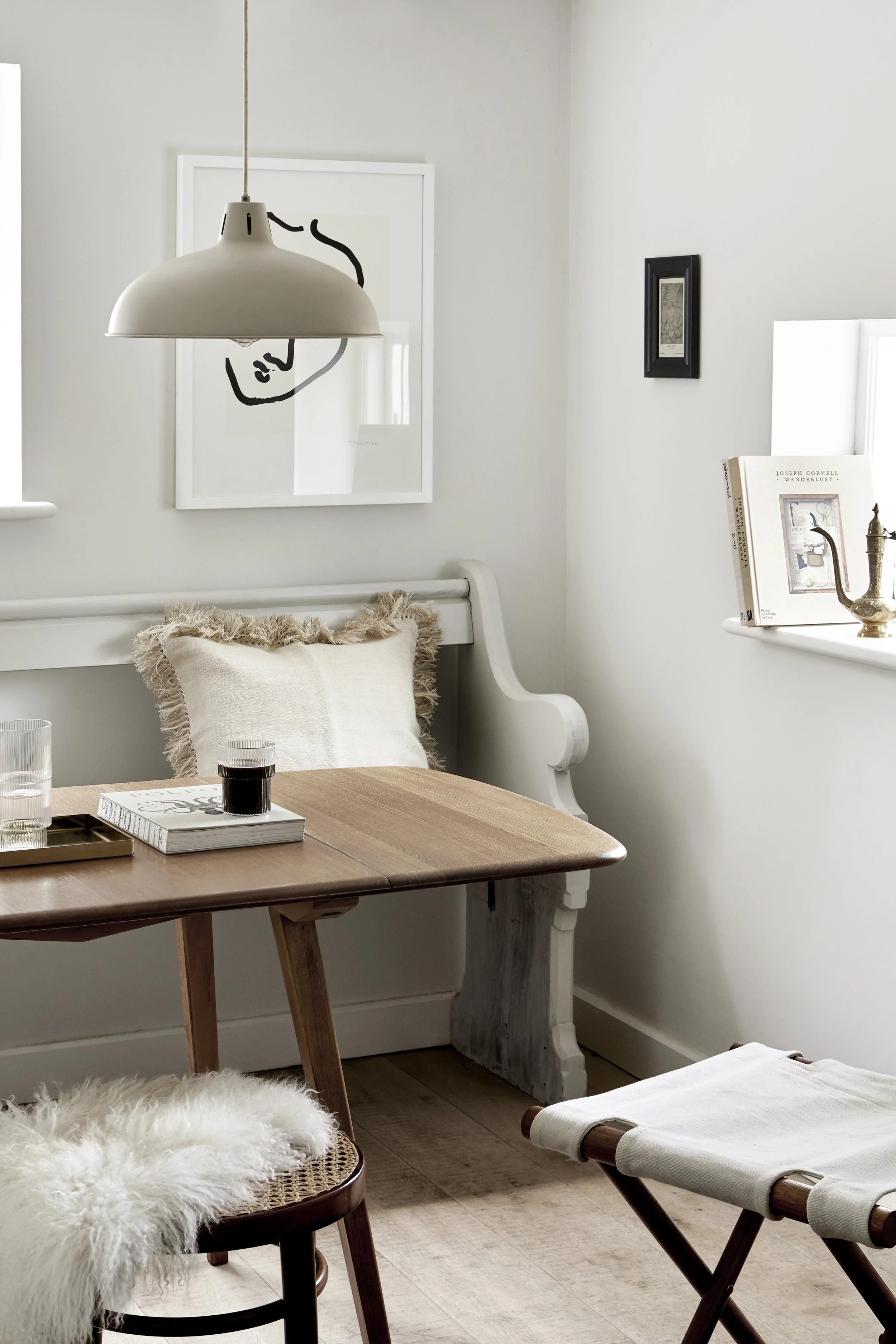 A cozy, minimalist dining nook with a wooden table, a vintage style white bench, and a wooden stool with a sheepskin cover. There is a white pillow on the bench, a glass of water, a book, and a small tray on the table. The room features white walls, 
