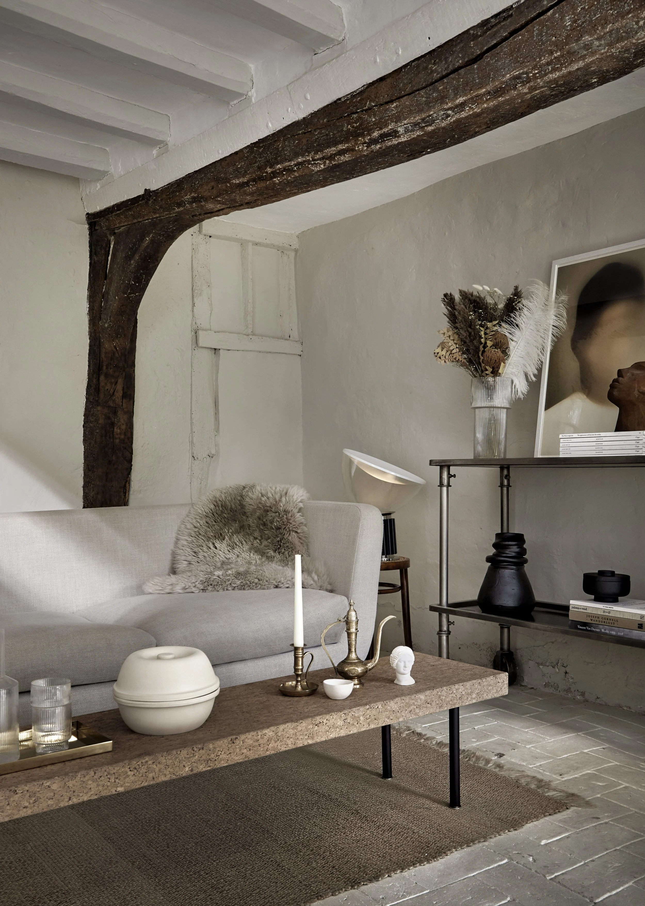Living room with a beige sofa, a coffee table with decorative items, a modern floor lamp, a shelving unit with vases and books, a large mirror, and a wall with a visible wooden beam and white textured wall.