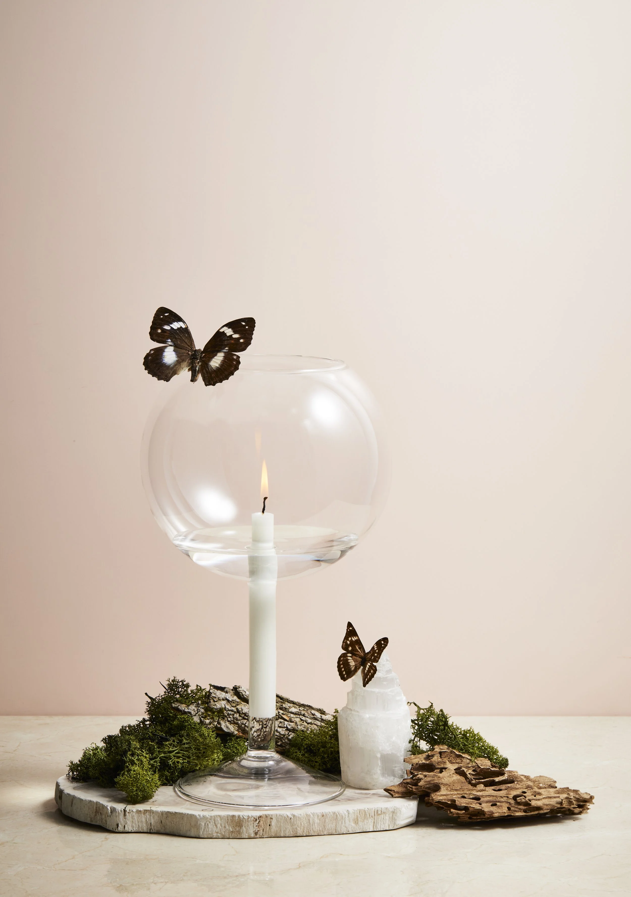 Decorative arrangement with a candle inside a glass bowl, surrounded by moss, a white mineral, bark, and two butterflies, one perched on the glass and the other on the mineral, on a round wooden base with a plain beige background.