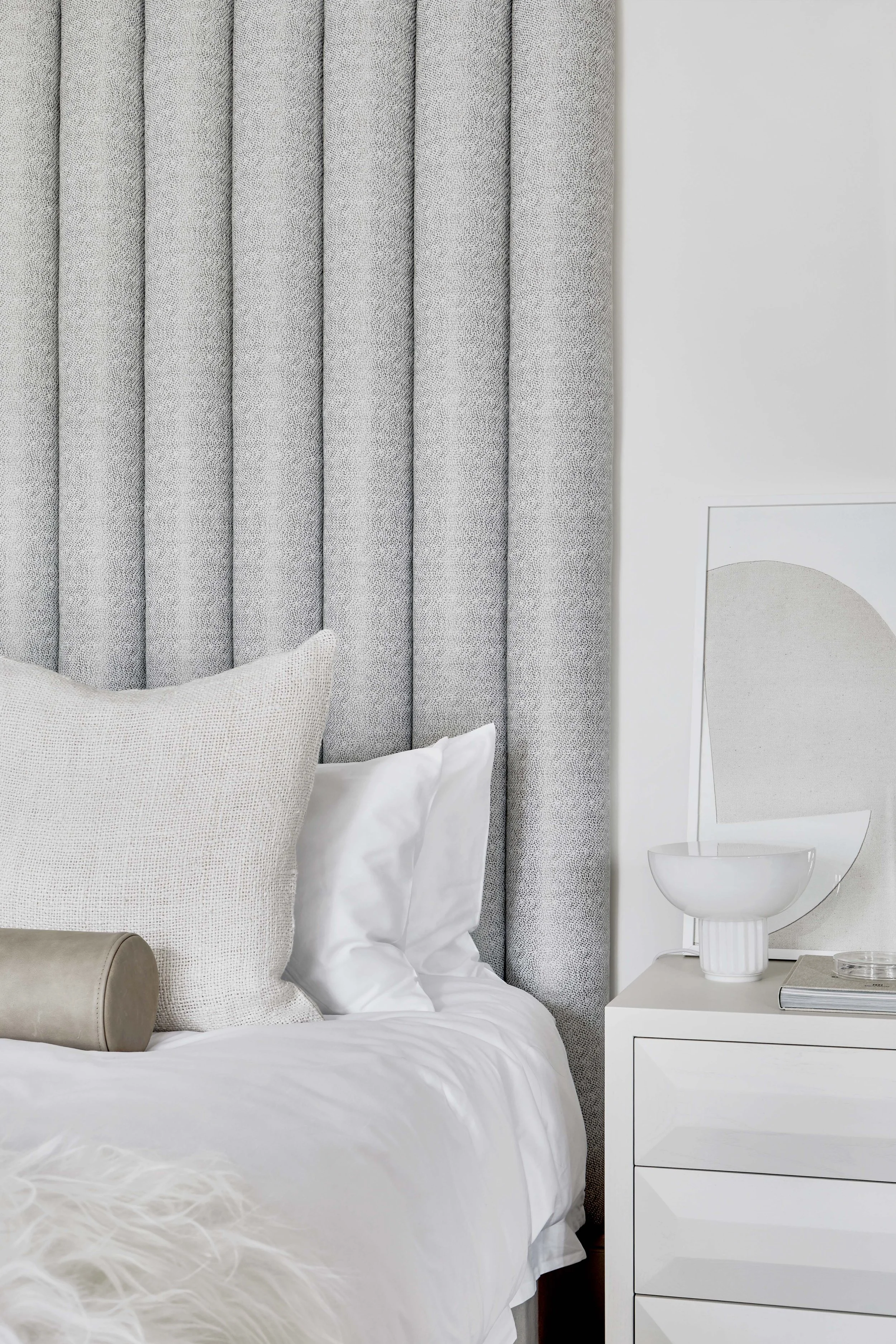 A bedroom with gray curtains, white bedding, decorative pillow, bedside white drawer, and modern art leaning against the wall.