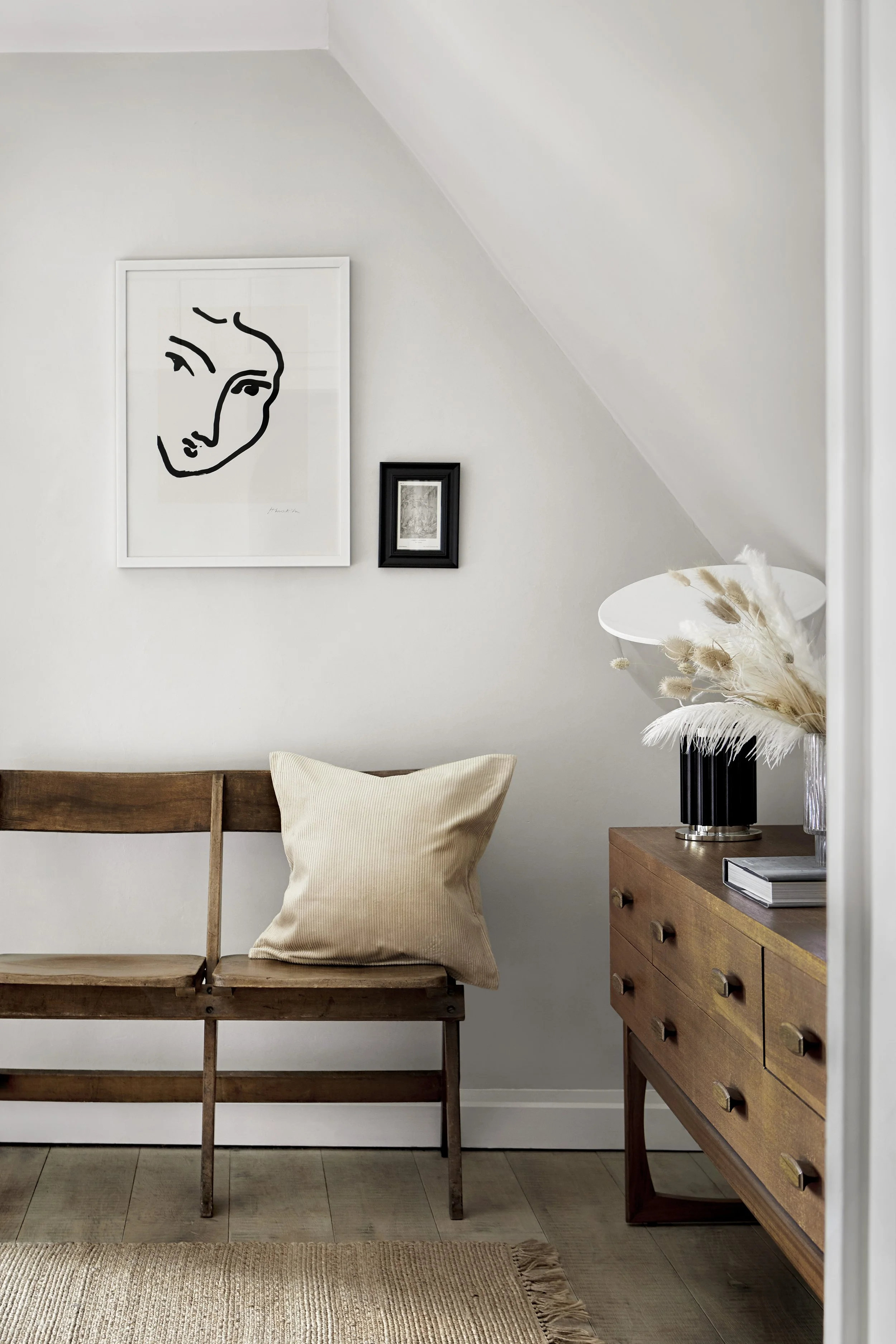 A cozy corner with a wooden bench, a beige pillow, a wooden sideboard with a black lamp, a vase with pampas grass, and framed art on a white wall.