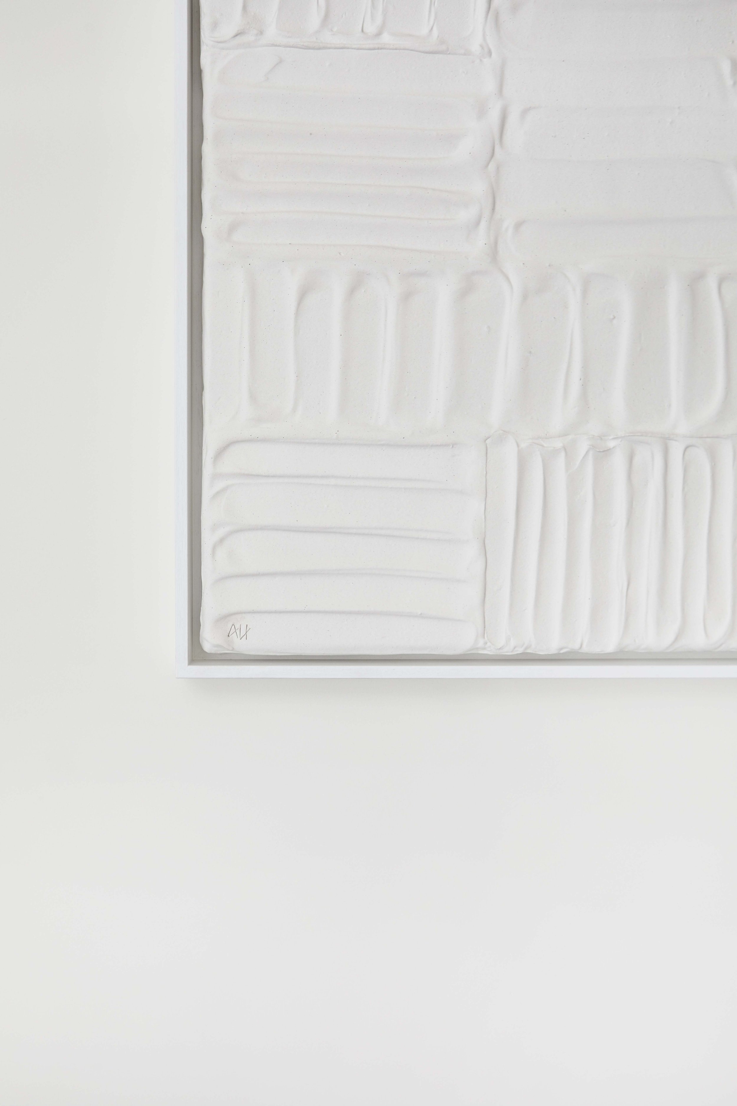 Close-up of a textured white abstract art piece with geometric patterns in a frame, on a white wall.