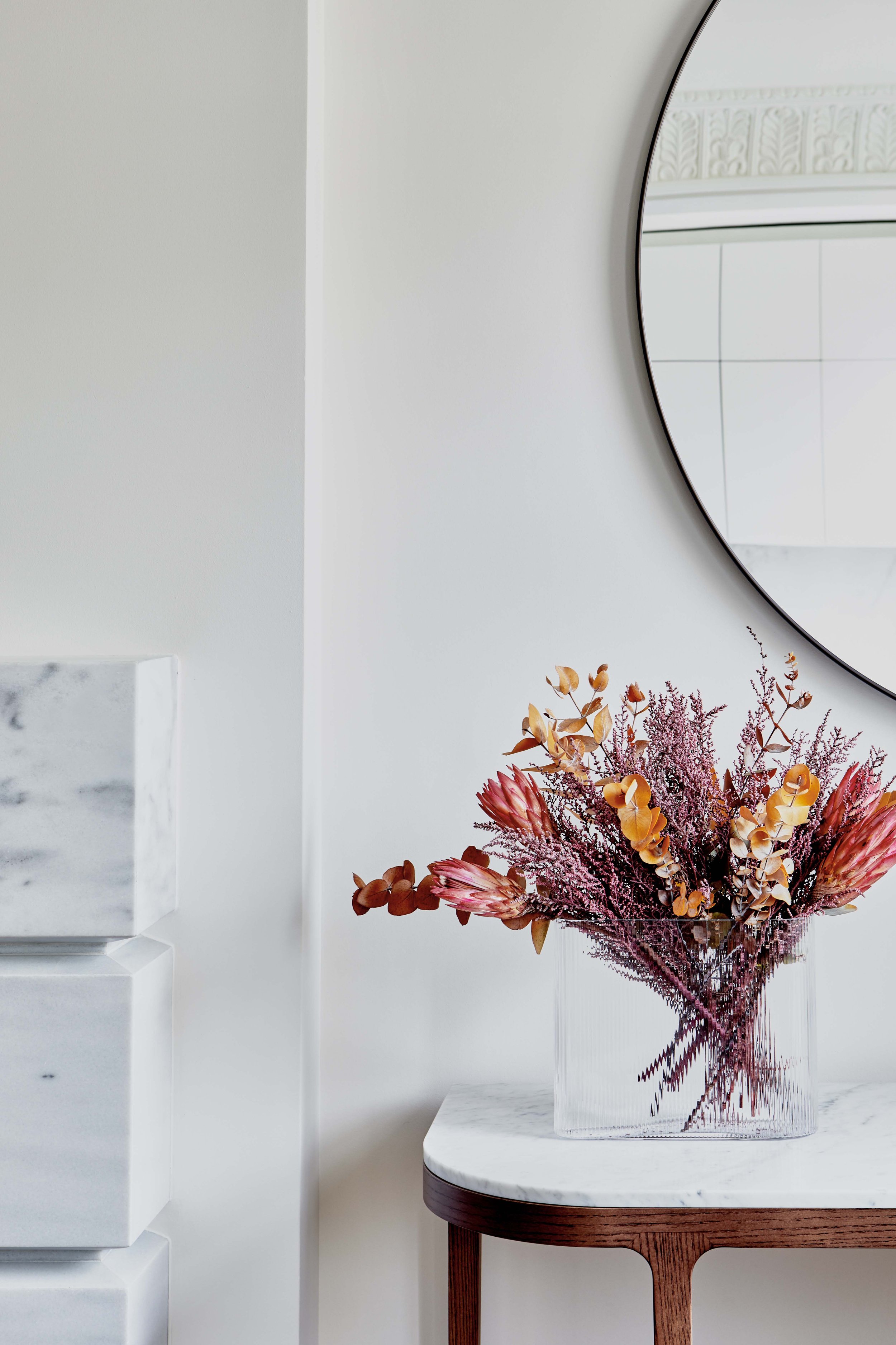 A clear glass vase filled with pink and purple dried flowers sits on a white marble-topped side table with a wooden leg, against a plain white wall with a round mirror reflecting part of a tiled wall.