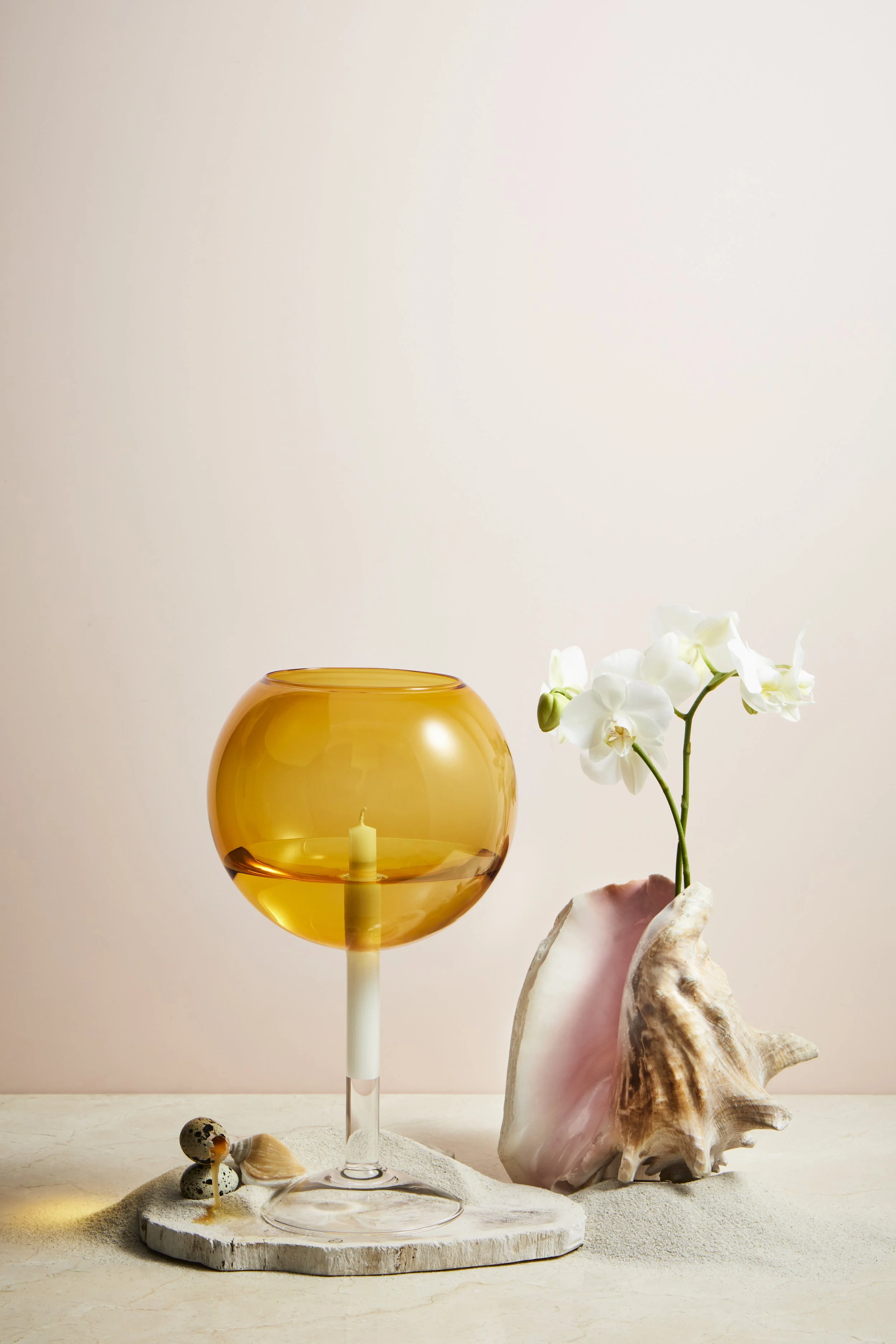 Decorative arrangement with a large amber glass bowl containing a white candle on a stand, a shell, and small mushrooms, next to a pink conch shell with white orchids growing out of it, on a light-colored surface with a plain beige background.