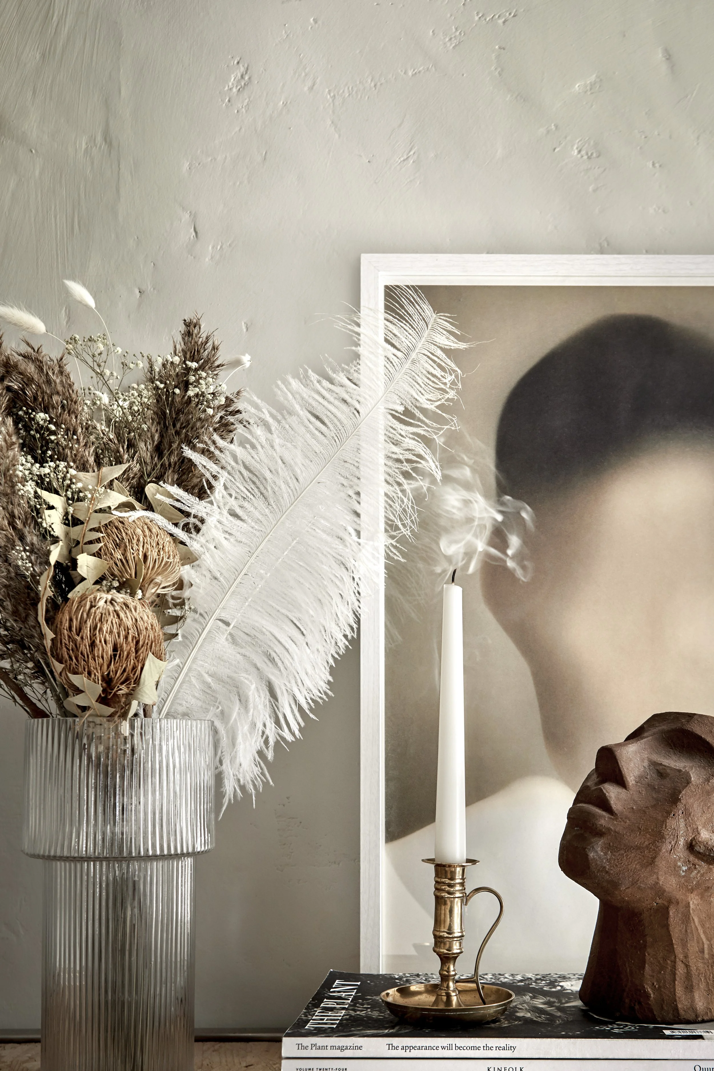 A decorative still life featuring a vase with dried flowers and grasses, a lit white candle in a brass holder, a large framed photograph of a woman with smoke near her face, and a wooden sculpture of a face on a table with magazines.