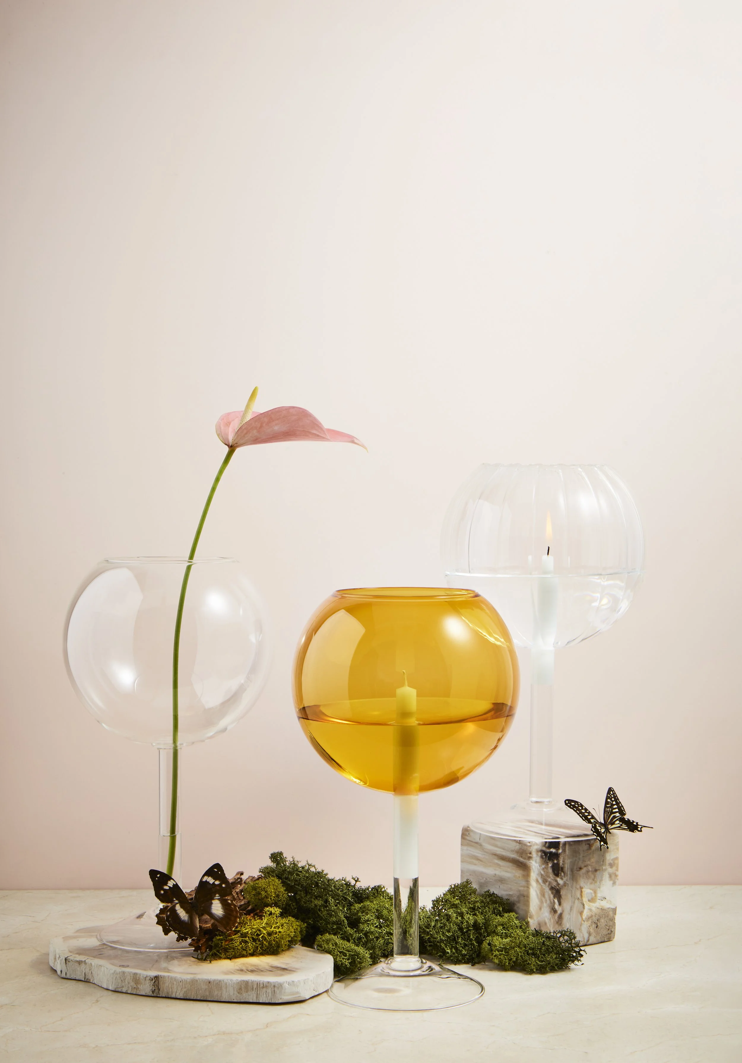 Decorative vases with candles and moss, butterflies, and a pink tulip on a light-colored surface against a neutral background.