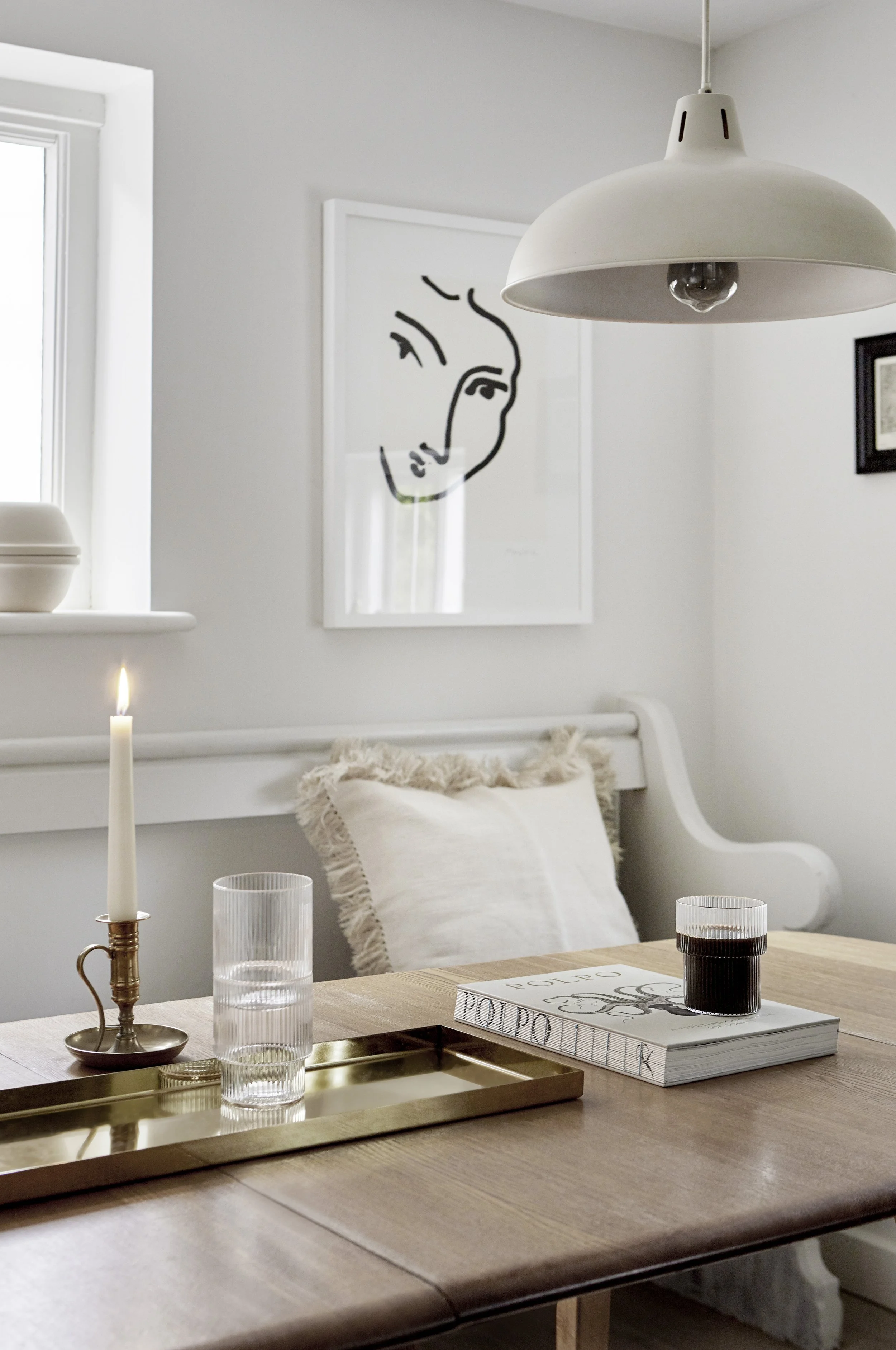 A dining room with a wooden table, a white pillow on a bench, a framed line drawing poster, a white ceiling lamp, and a window.