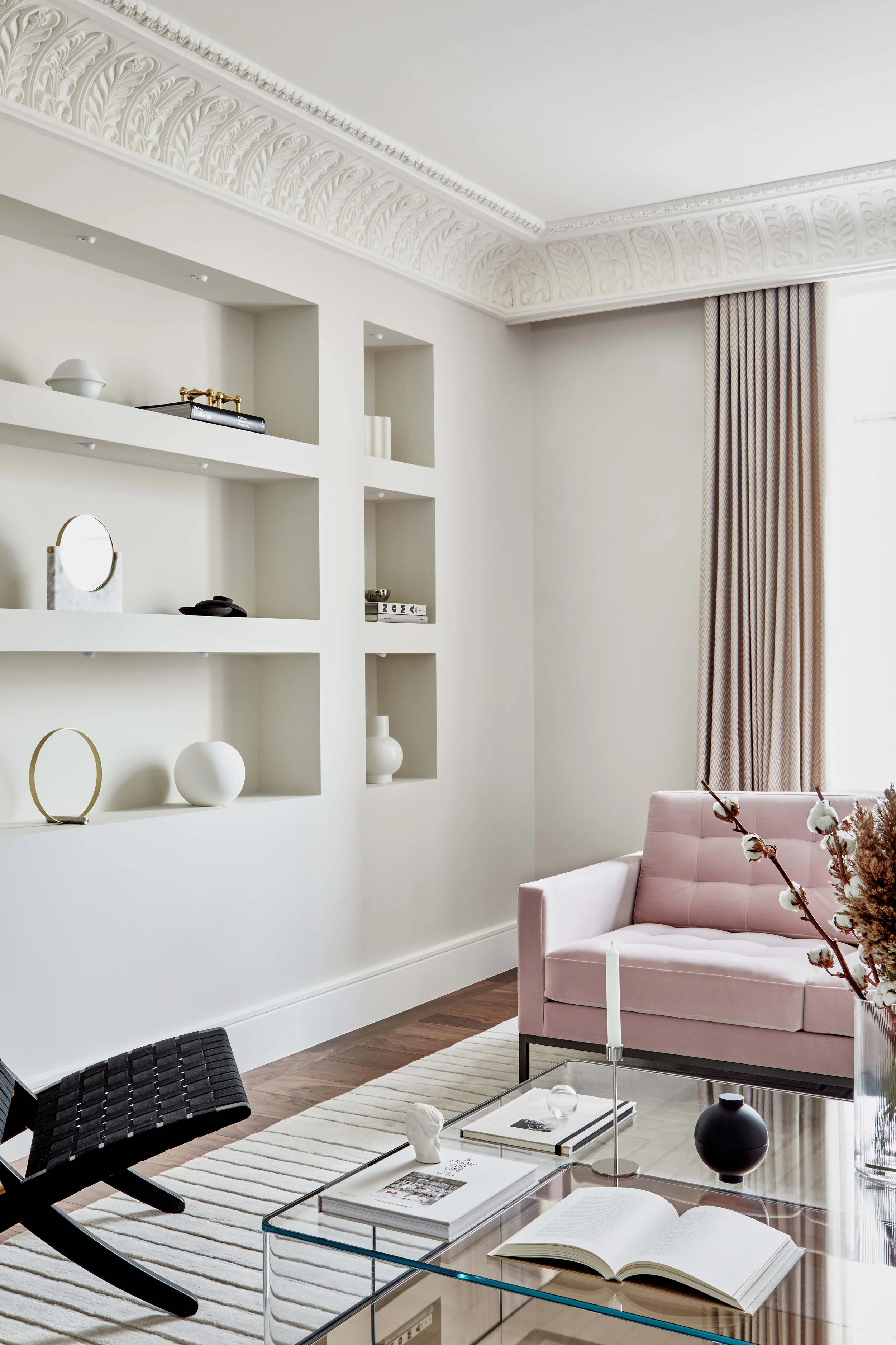 Living room with white walls, built-in shelves filled with decorative items, light pink sofa, glass coffee table, striped rug, and cream curtains.
