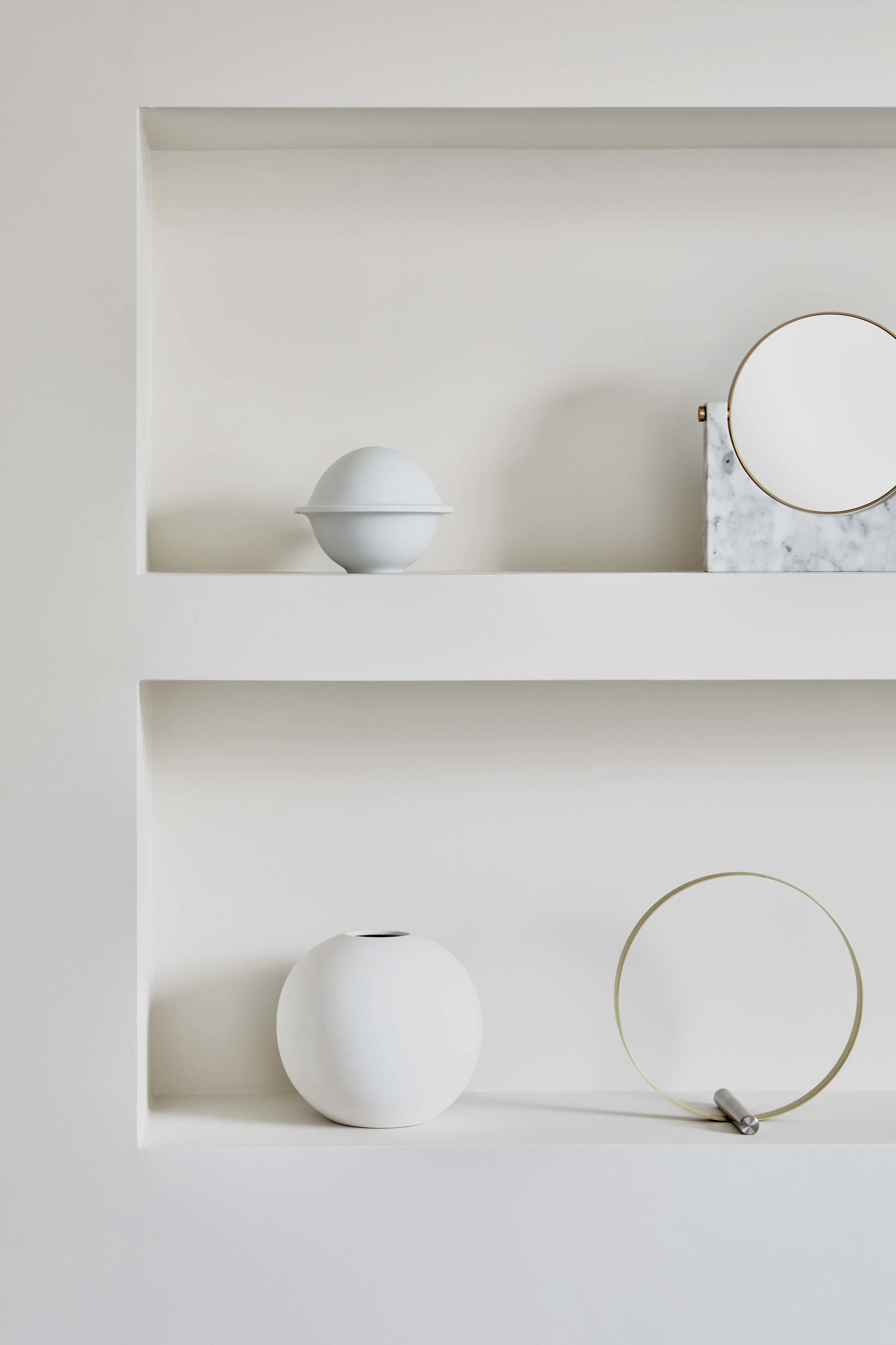 Minimalist display of white decor items on built-in wall shelves, including a round vessel, a mirror with a marble base, a white spherical vase, and a gold metal circle