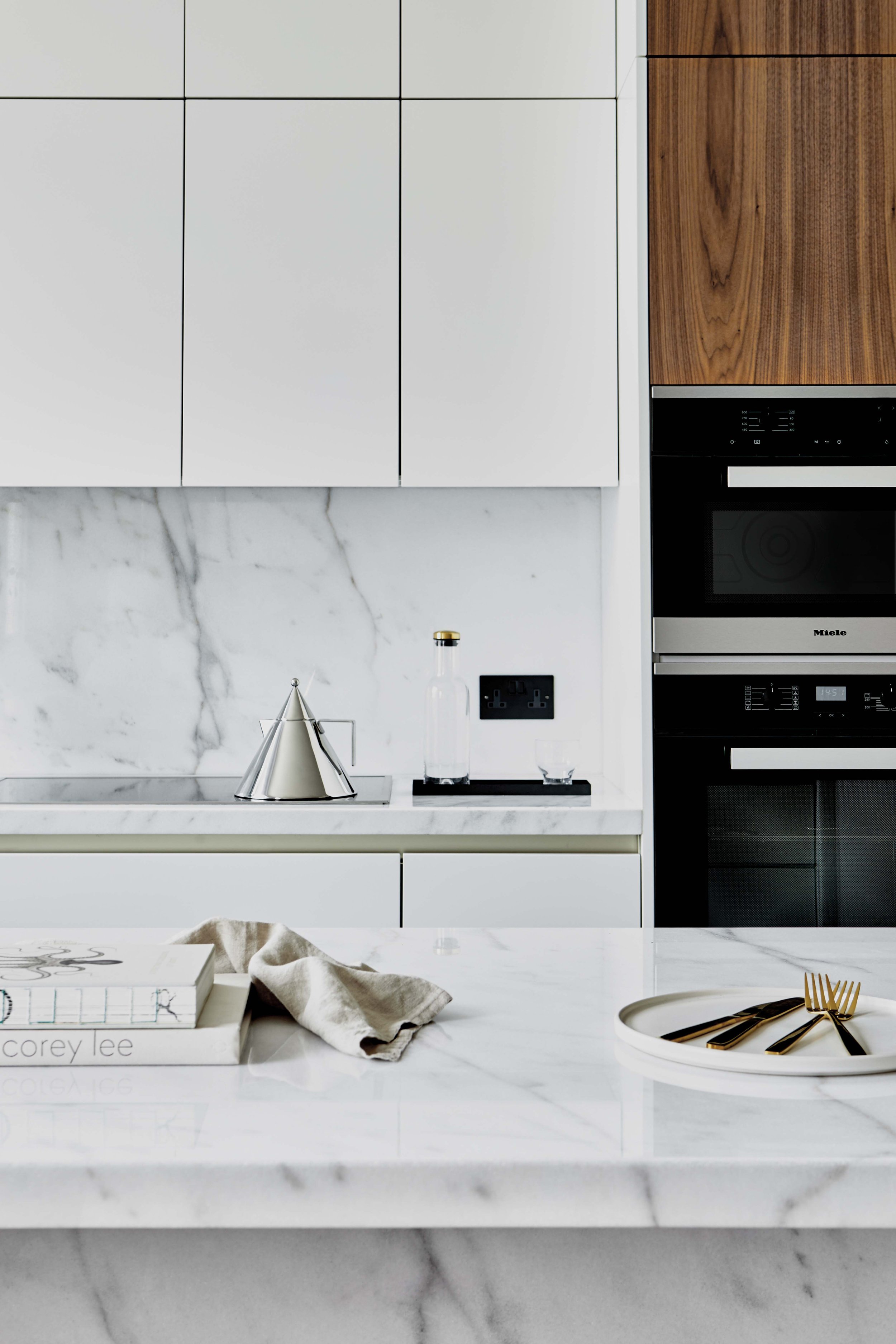 Modern kitchen with white cabinets, marble backsplash, and a marble countertop. Items include a silver kettle, a glass bottle, a small glass, a book, a cloth, and a tray with gold utensils.