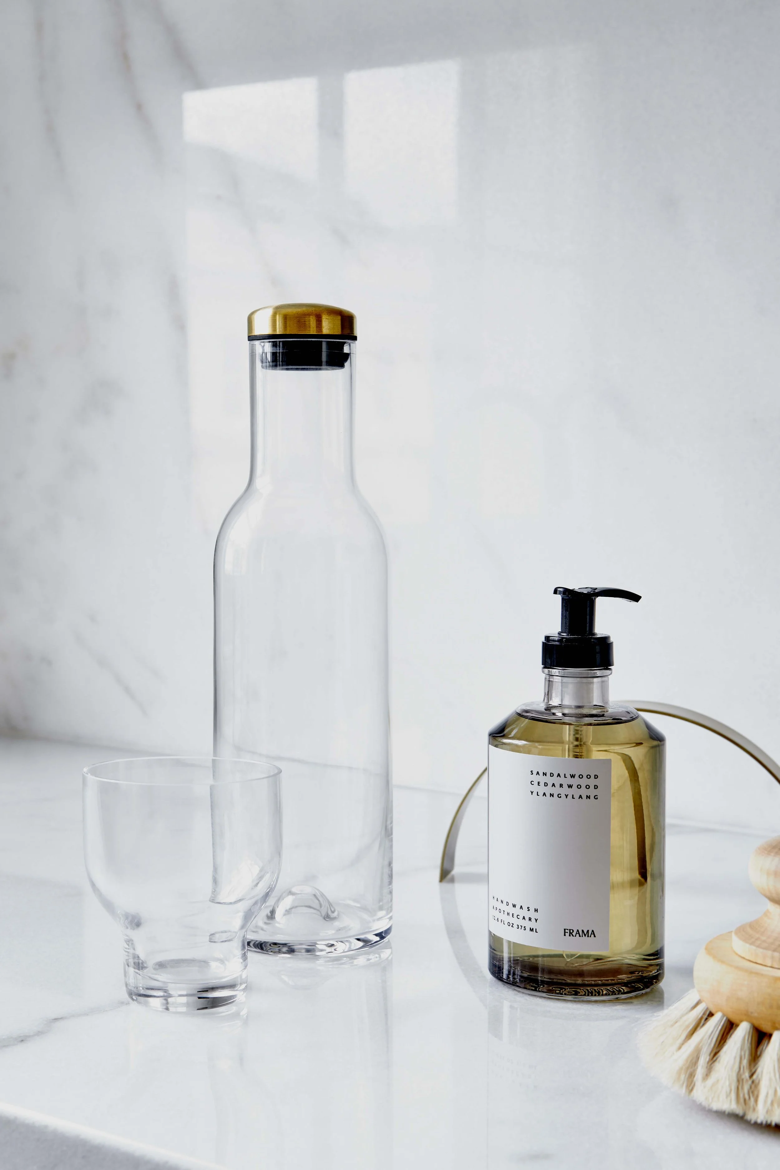 Empty glass water bottle with a gold lid, a glass tumbler, and a bottle of hand soap on a white marble surface.