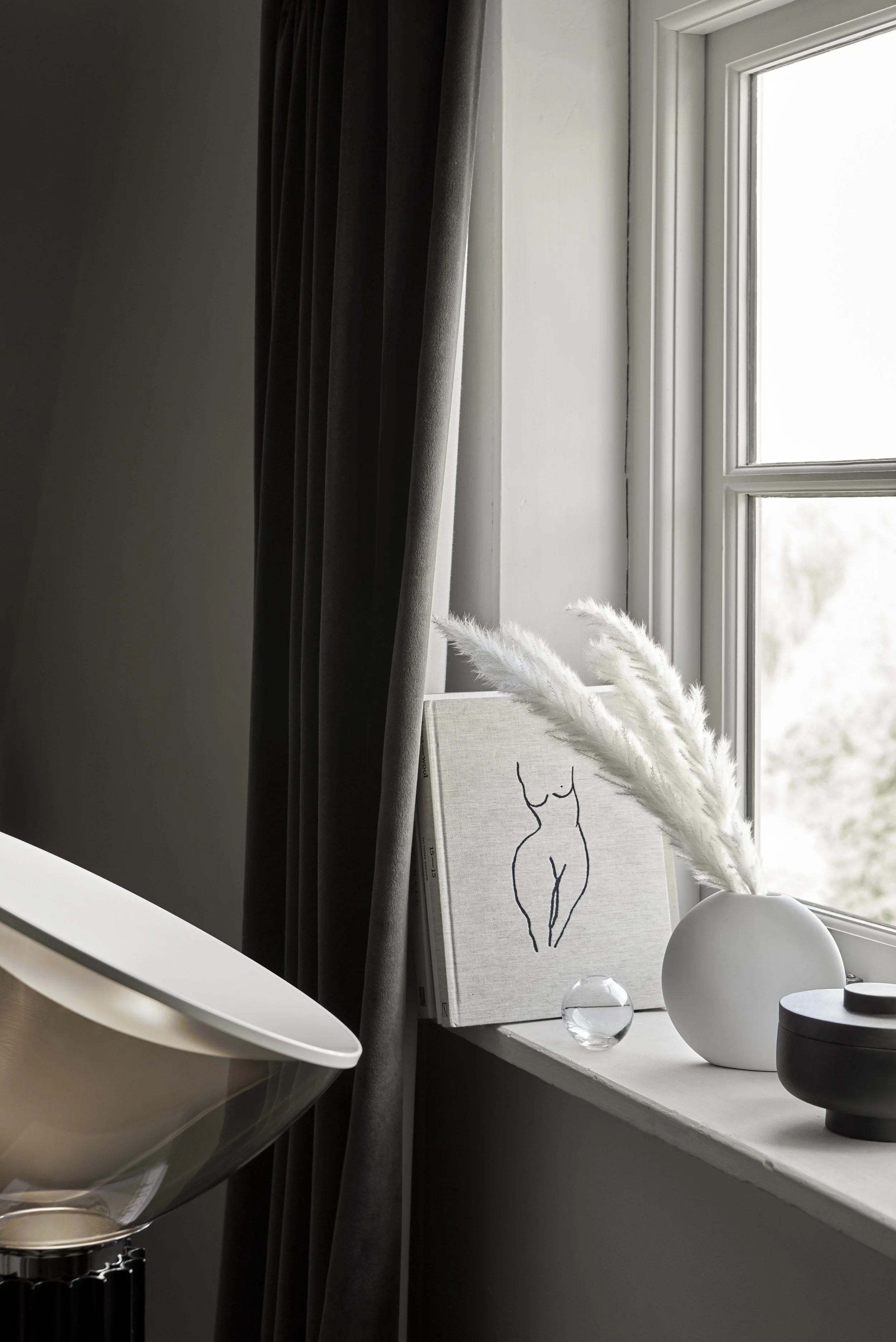 Decorative interior corner with a sketch art book, vase with white pampas grass, small glass sphere, and black container on a window sill beside a large window with dark curtains.